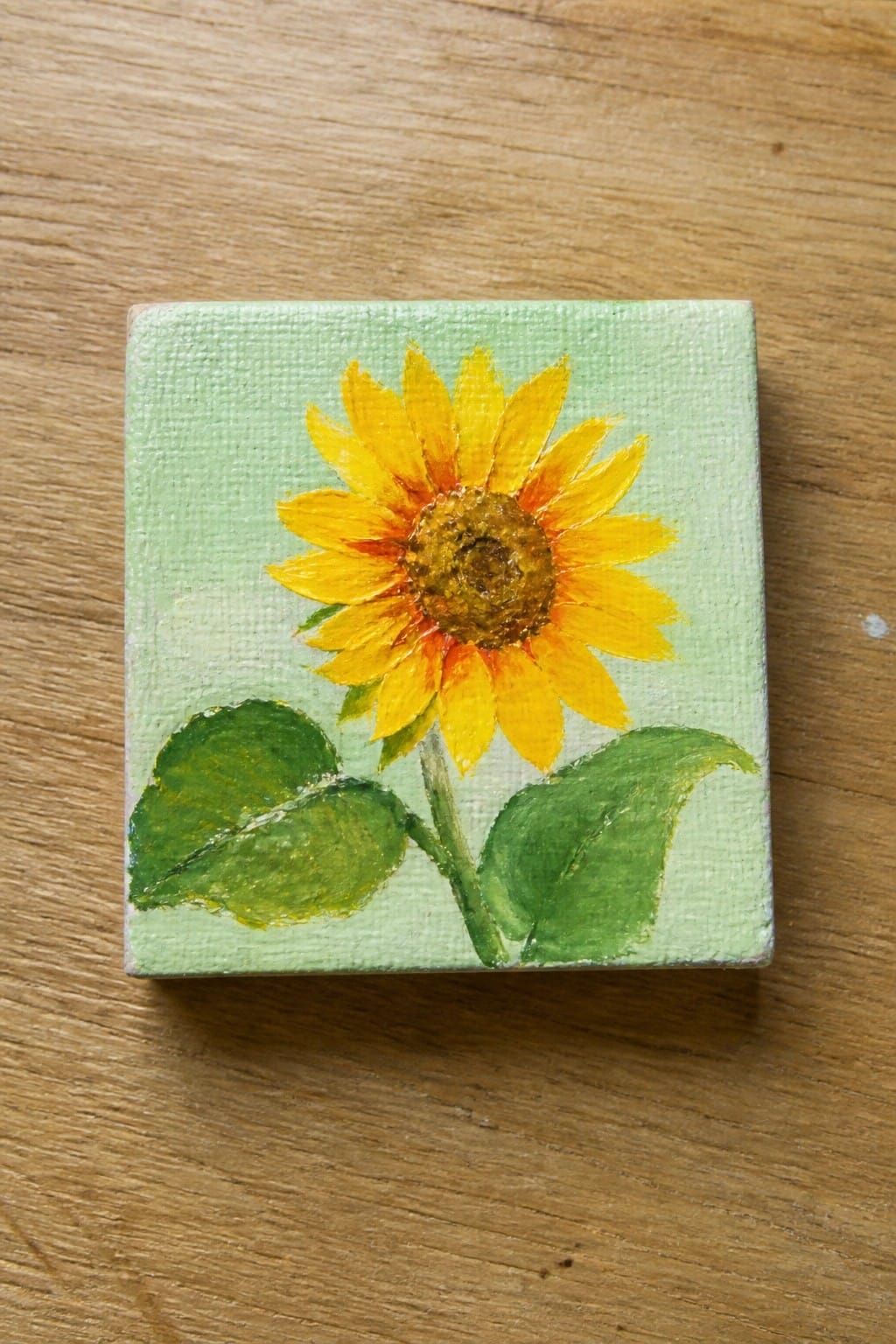 Small square canvas with yellow sunflower, green leaves on light green background atop wood