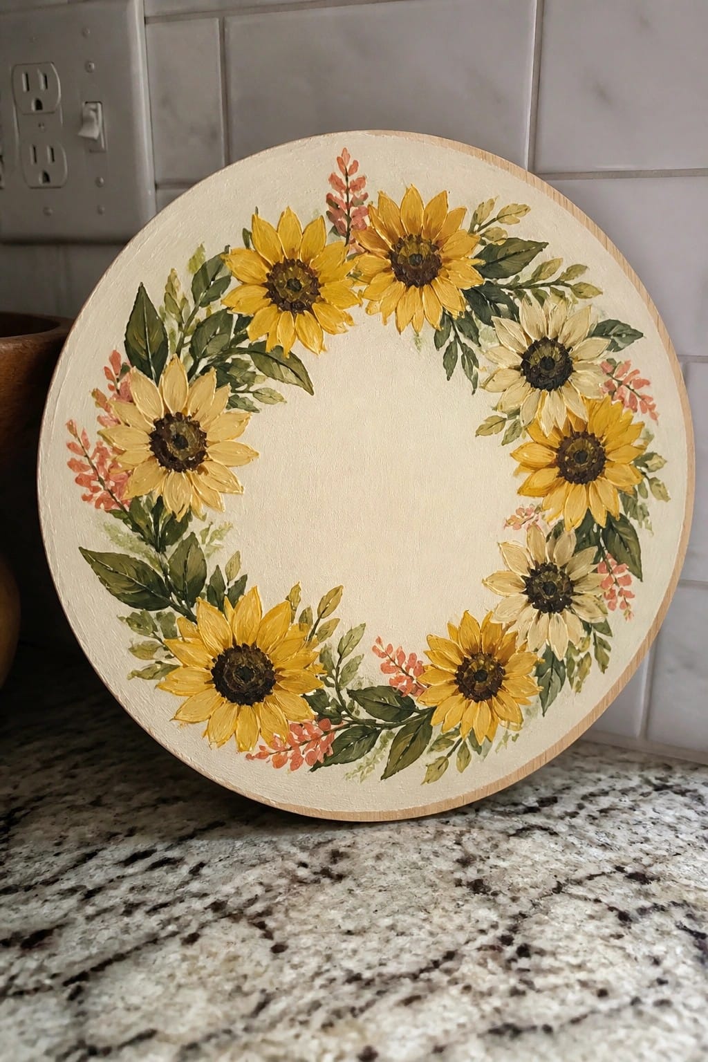 A round wooden board with a painted wreath of yellow sunflowers, green leaves, and small red accents on a cream background.