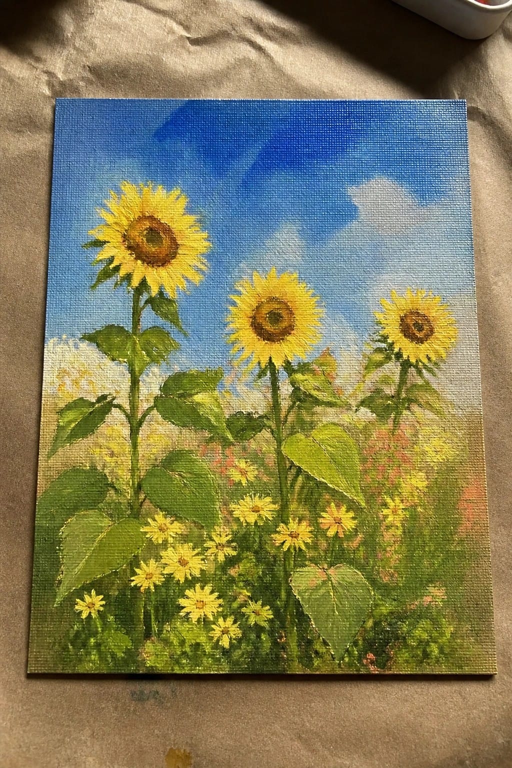 Three yellow sunflowers with green leaves under a bright blue sky with clouds and wildflowers
