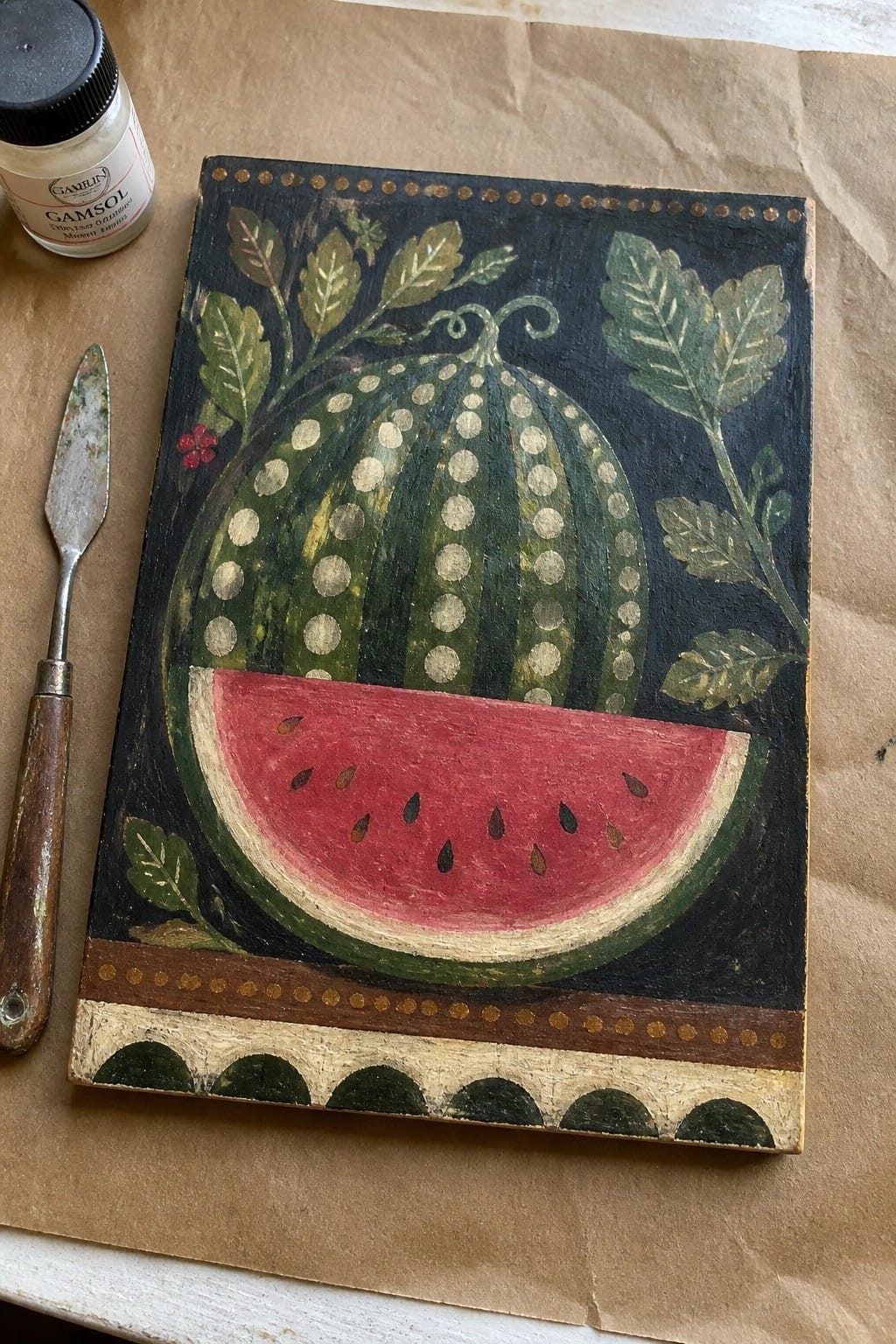 Folk art watermelon painting with red slice and leaves beside palette knife and jar.