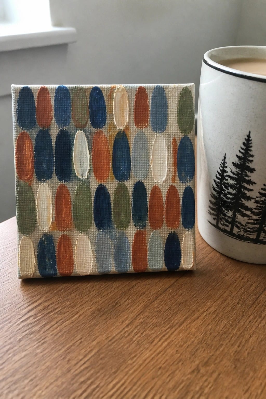 Small canvas with colorful oval pattern beside white mug with tree design on wood
