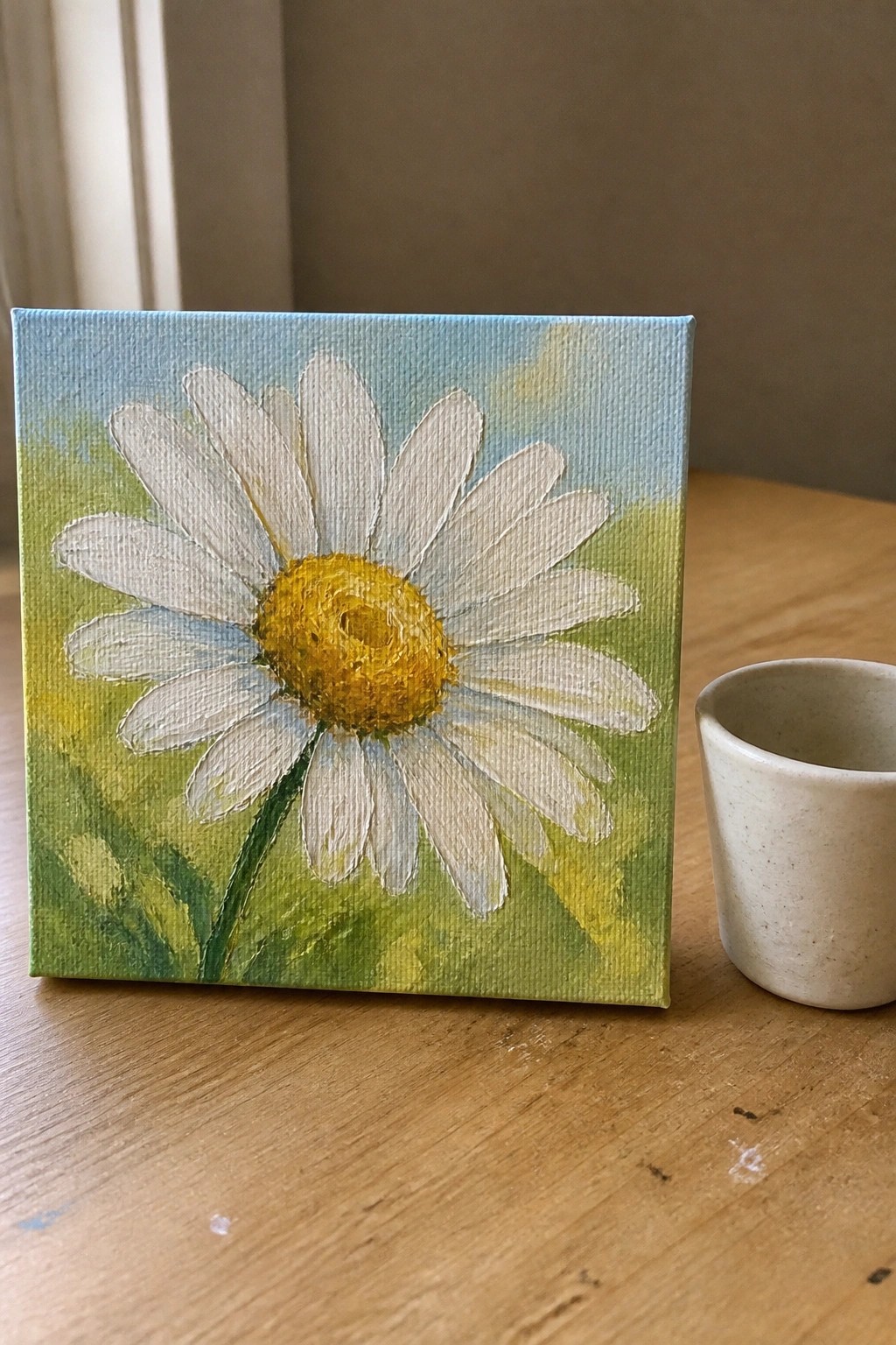 Canvas painting of white daisy with yellow center beside small ceramic cup on wood