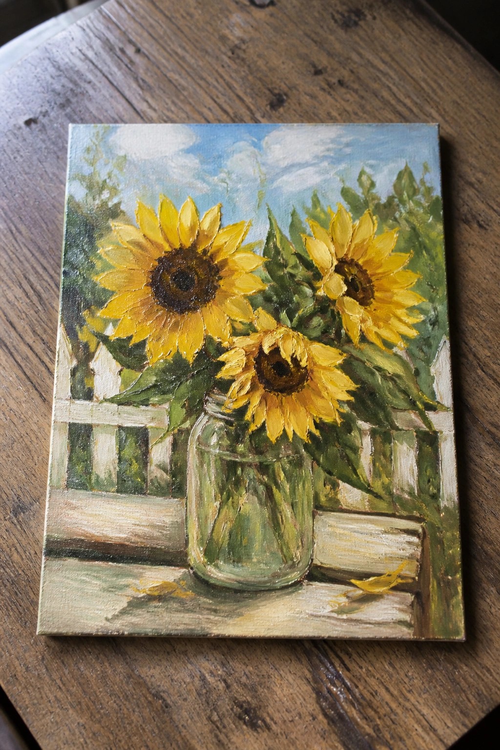 Three sunflowers in a glass jar on wood with white fence and blue sky behind.