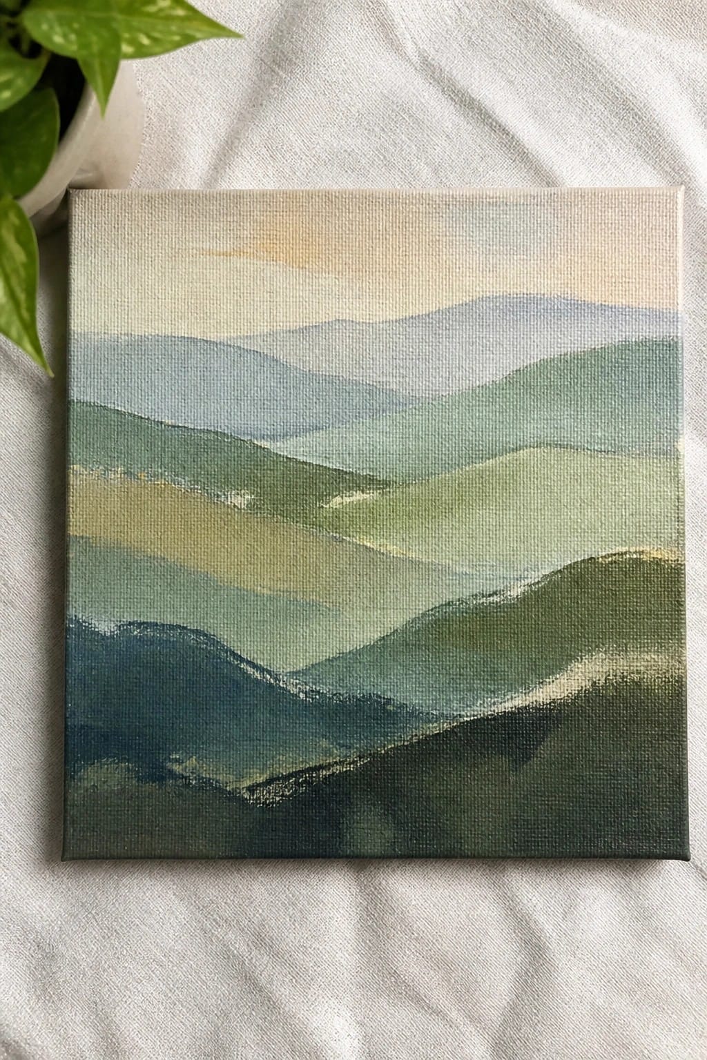 Abstract landscape painting of layered green blue hills on white fabric beside potted plant