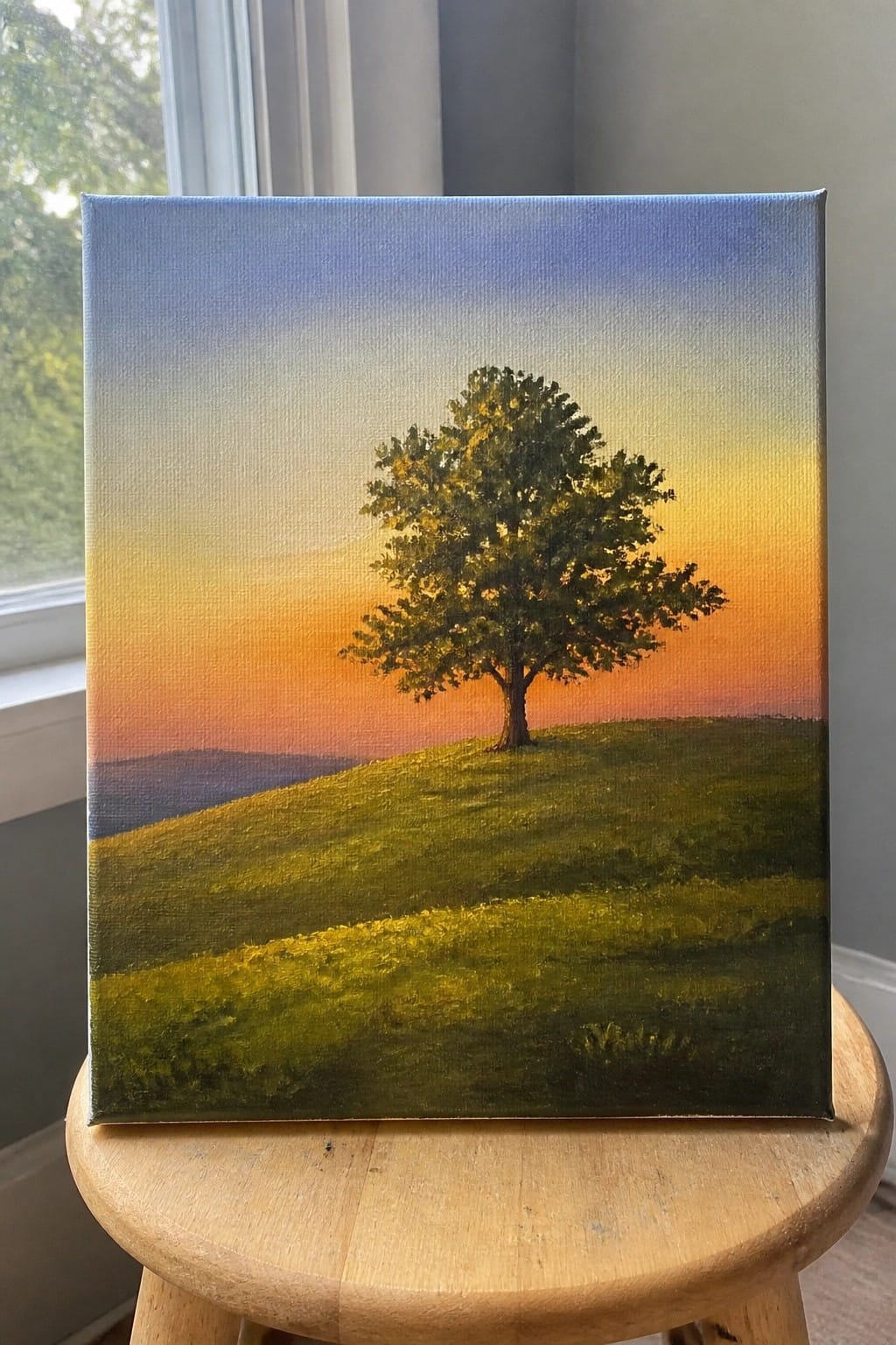 An oil painting of a single tree on a green hill under a blue to orange sunset sky with distant hills.