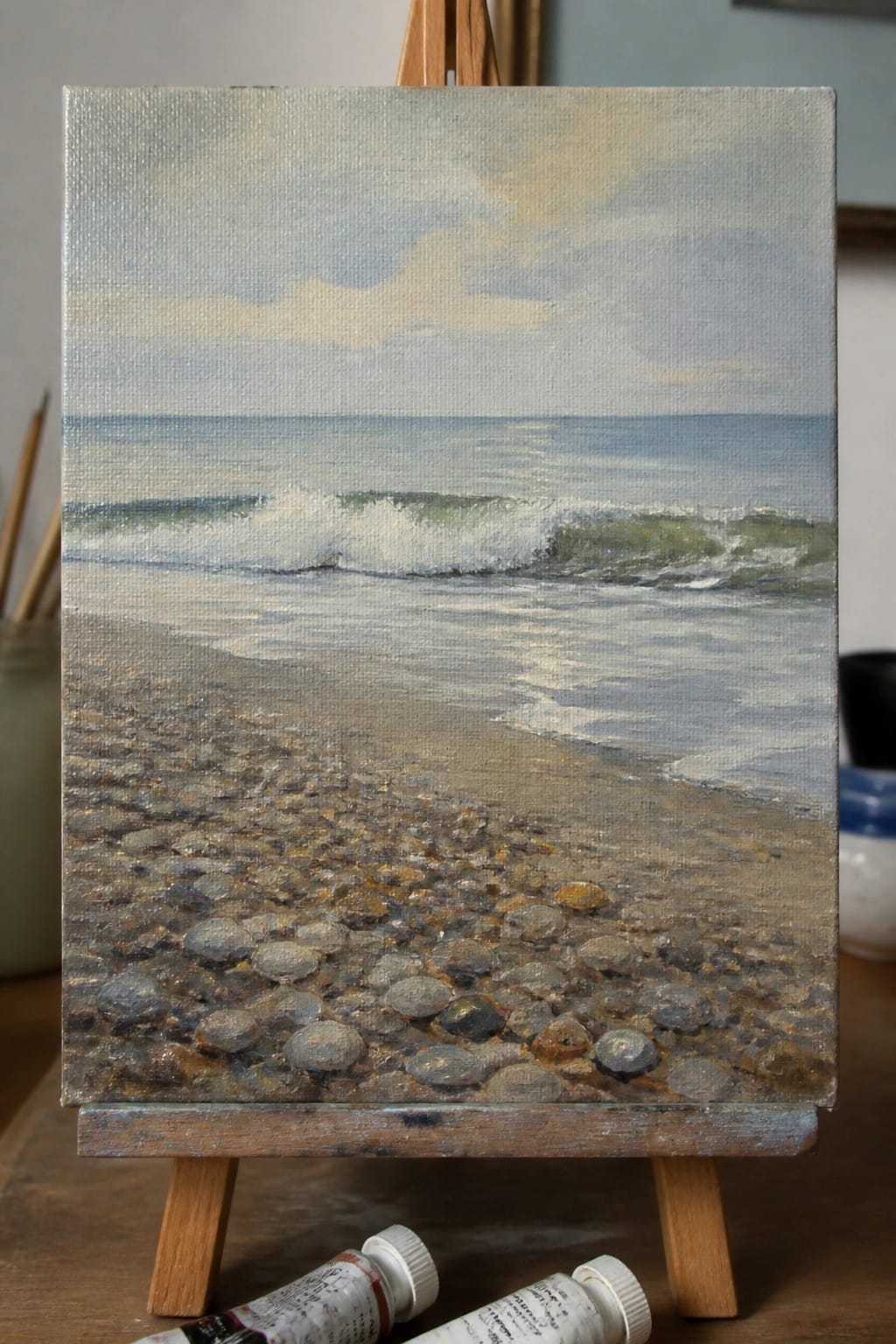 Oil painting of pebble beach with waves on easel, paint tubes below