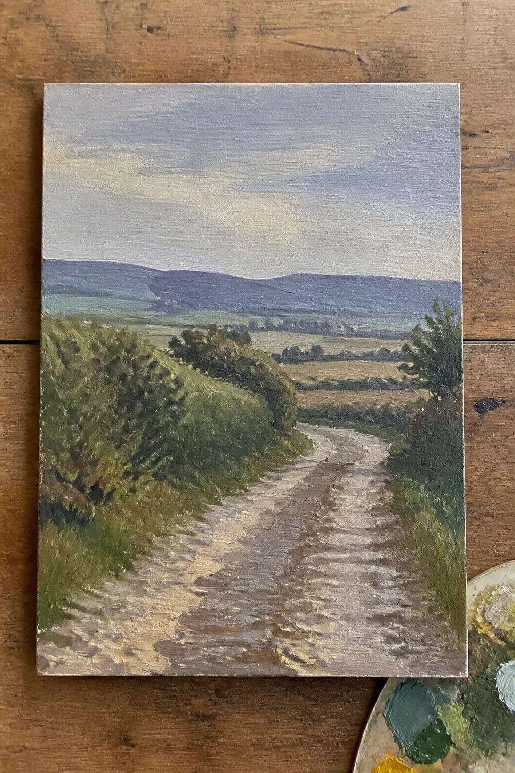 Oil painting of winding dirt path through green hills and fields under blue sky