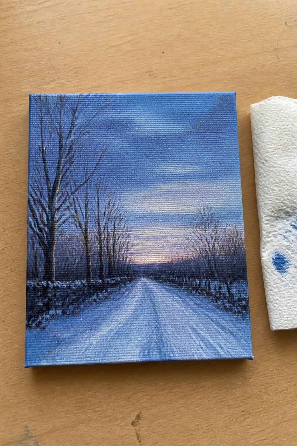Small canvas painting of snowy road with bare trees at dusk beside blue paint towel on wood.