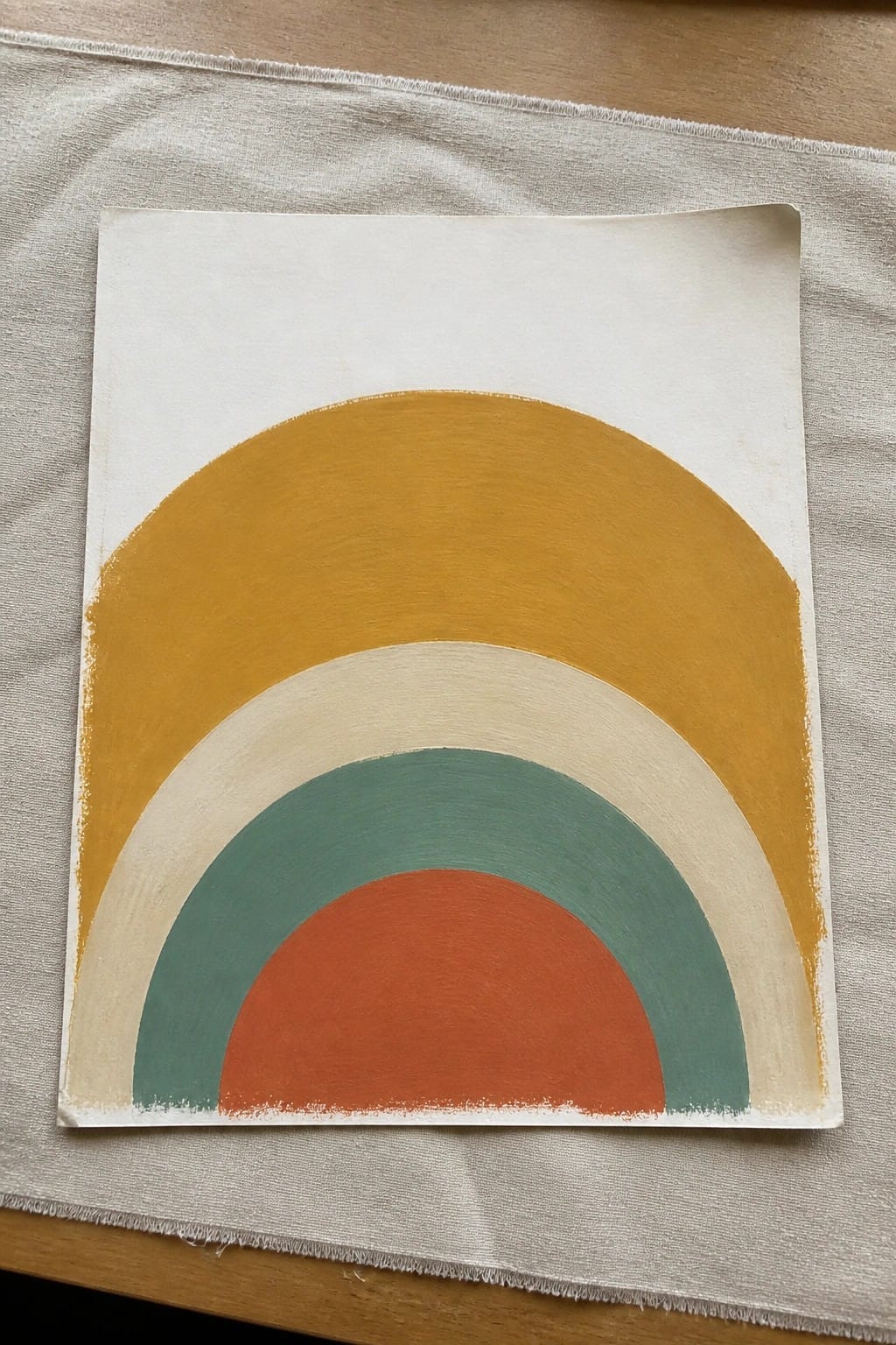 Rainbow painting with yellow, beige, teal, and orange arcs on white paper over fabric.