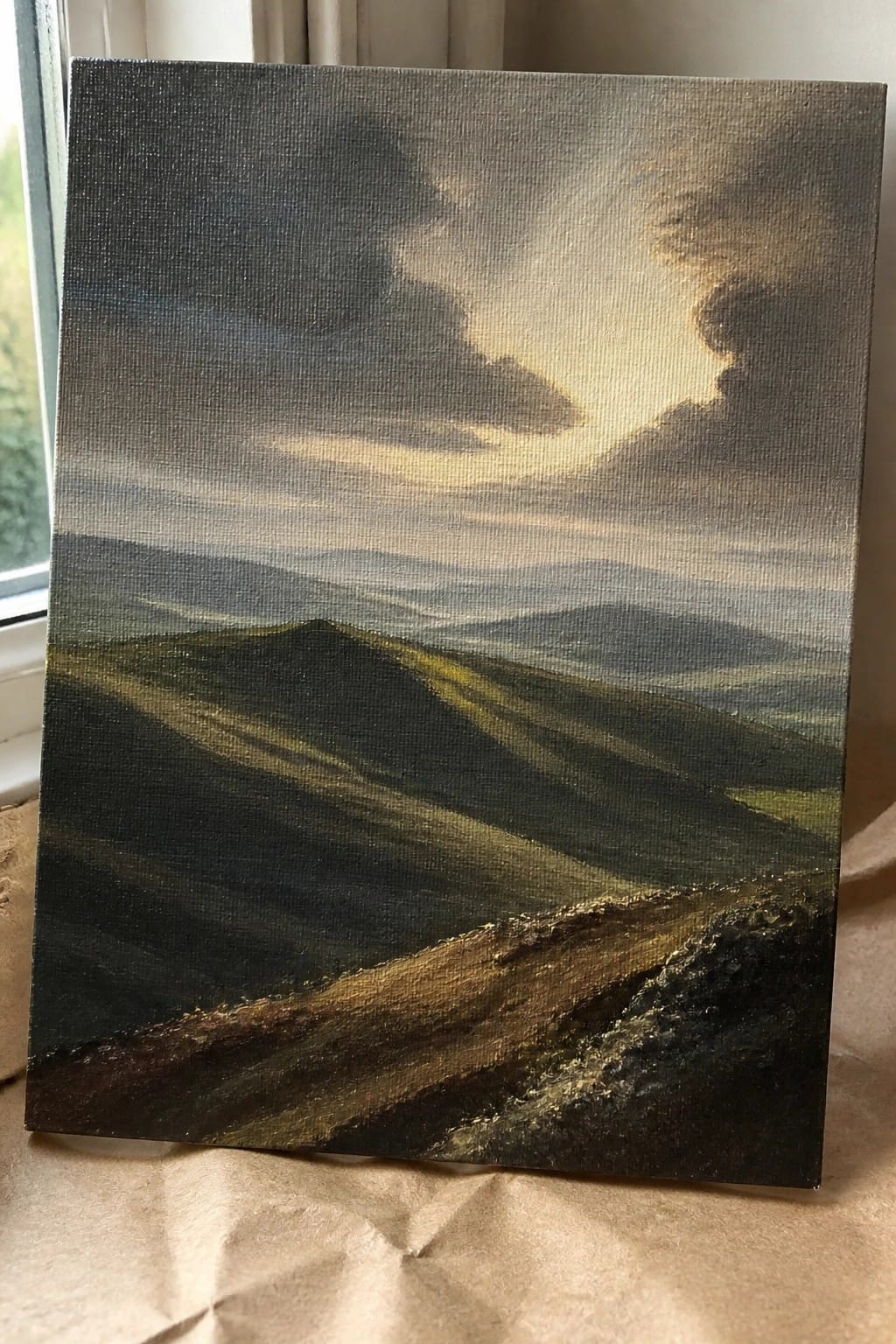 Dark landscape painting of rolling hills beneath stormy clouds with golden sunlight rays.