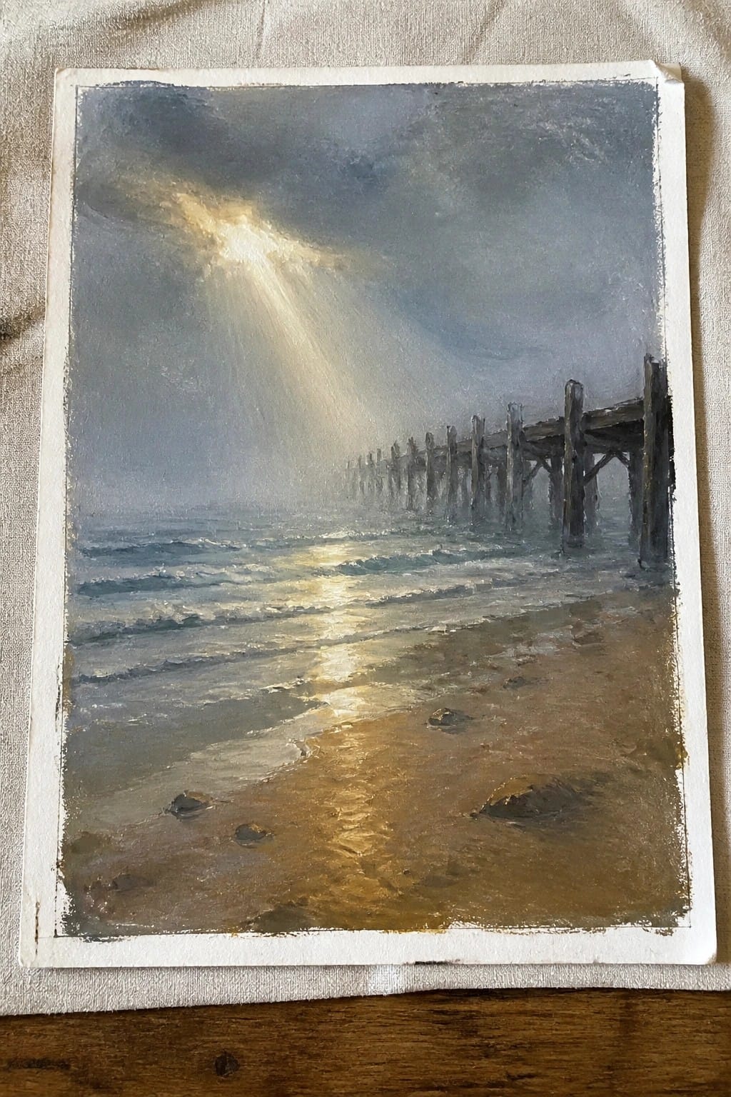Dramatic oil painting of sunlight piercing clouds over waves, sandy beach, and ruined pier.