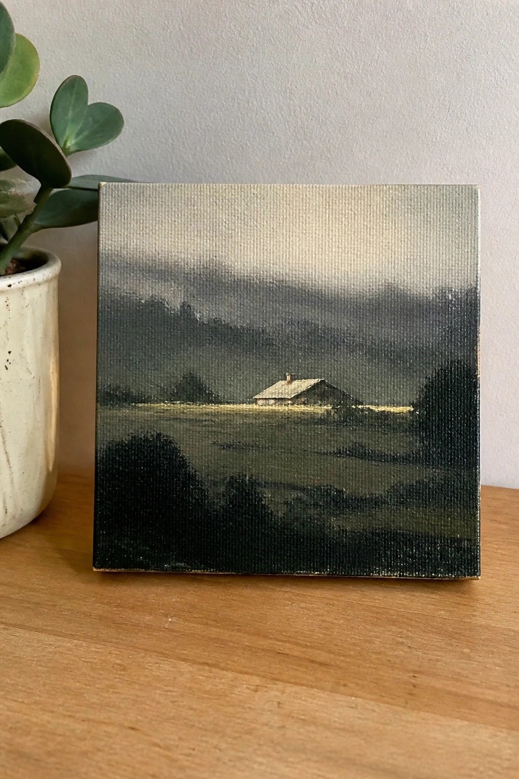 Small canvas landscape painting of house under clouds beside potted plant on wood