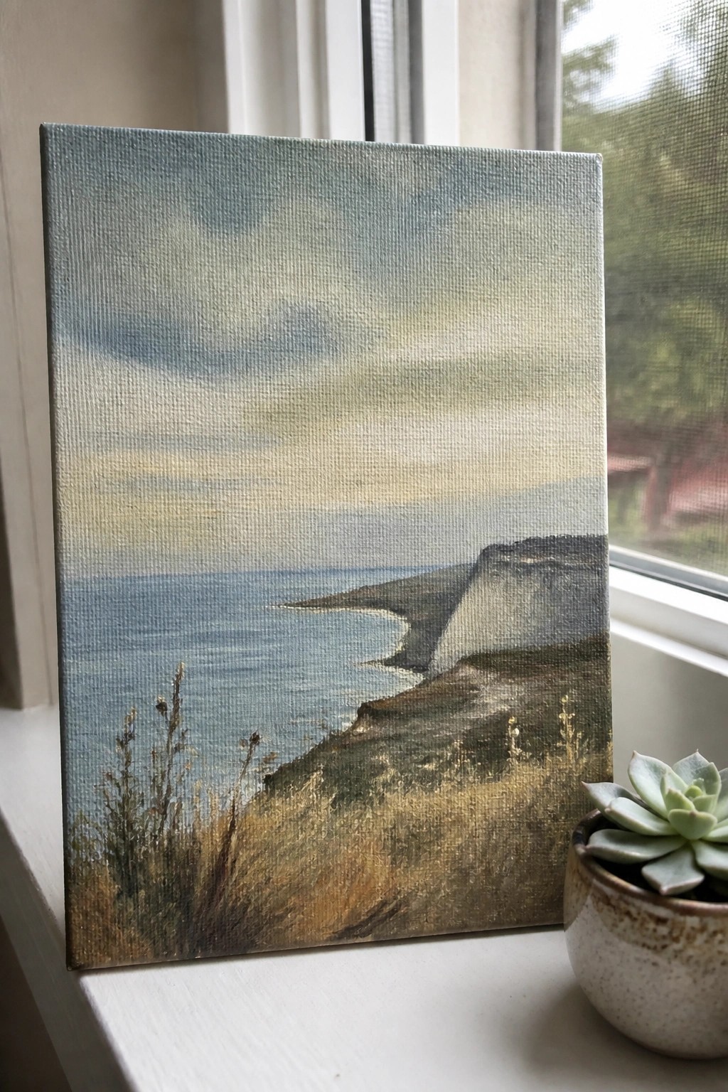 Seascape painting of white cliffs and ocean on windowsill beside potted succulent.