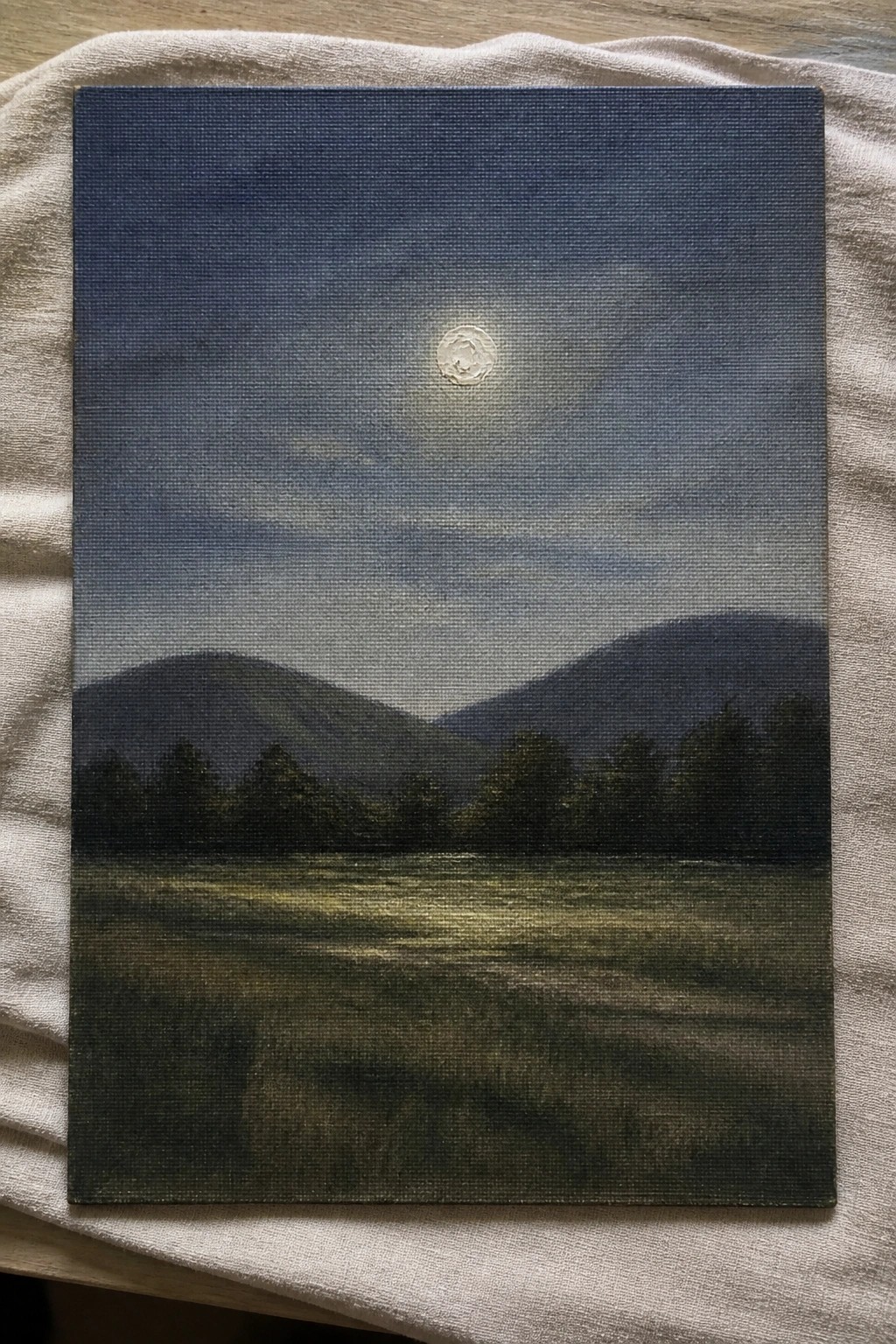Painting on canvas showing full moon above dark mountains and grassy field on beige cloth.