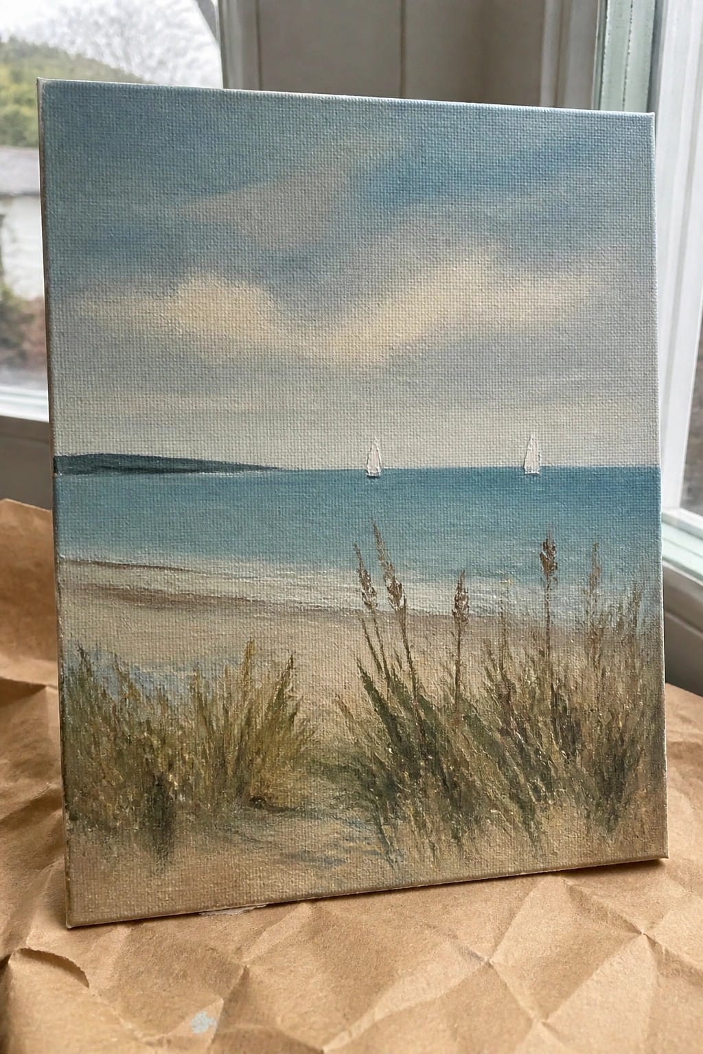 Seascape painting on canvas with blue ocean, two sailboats, sky, and beach grasses in foreground.