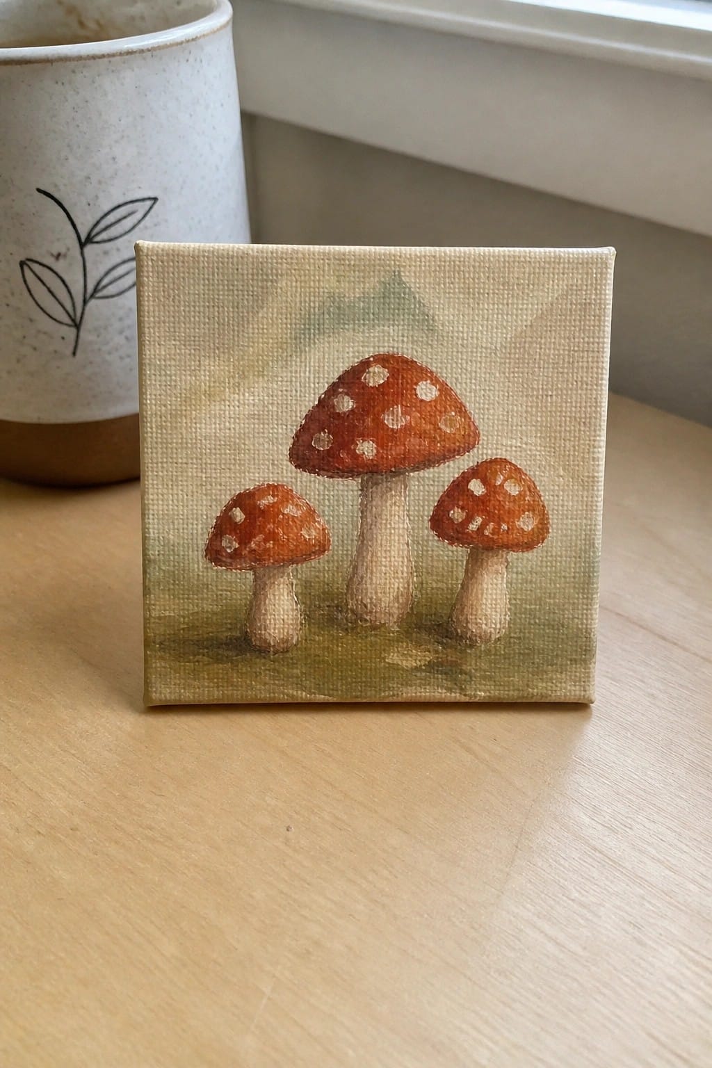 Small canvas painting of three red-spotted mushrooms beside a white mug on wood