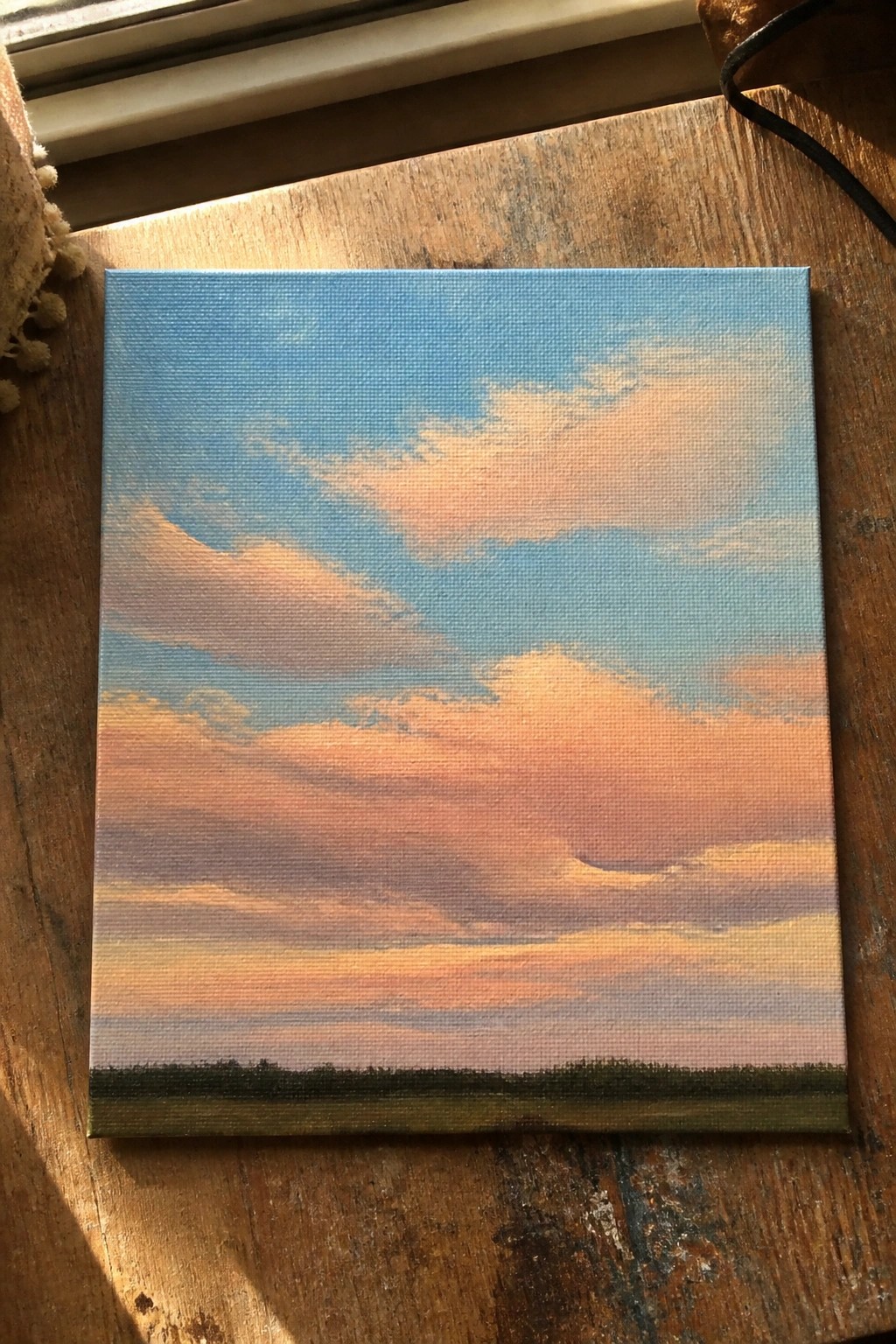 Small canvas painting of sunset sky with orange-pink clouds above green horizon on wood.