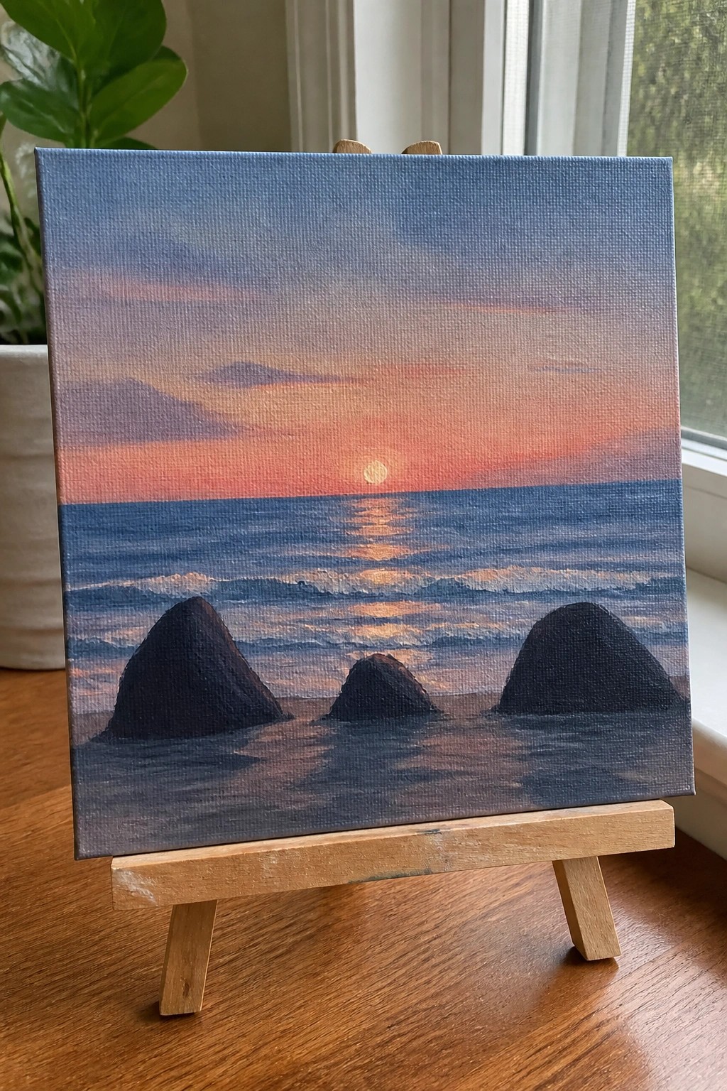 Canvas painting on easel of sunset over ocean with three dark rocks