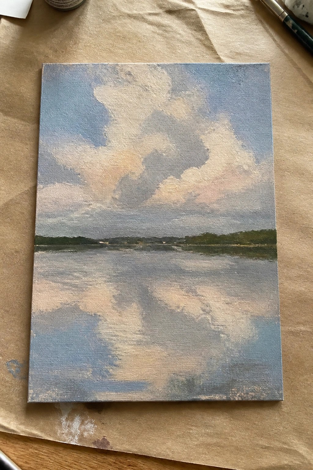 An oil painting of calm water reflecting a sky with soft blue, gray, and pink clouds above a distant green shoreline.