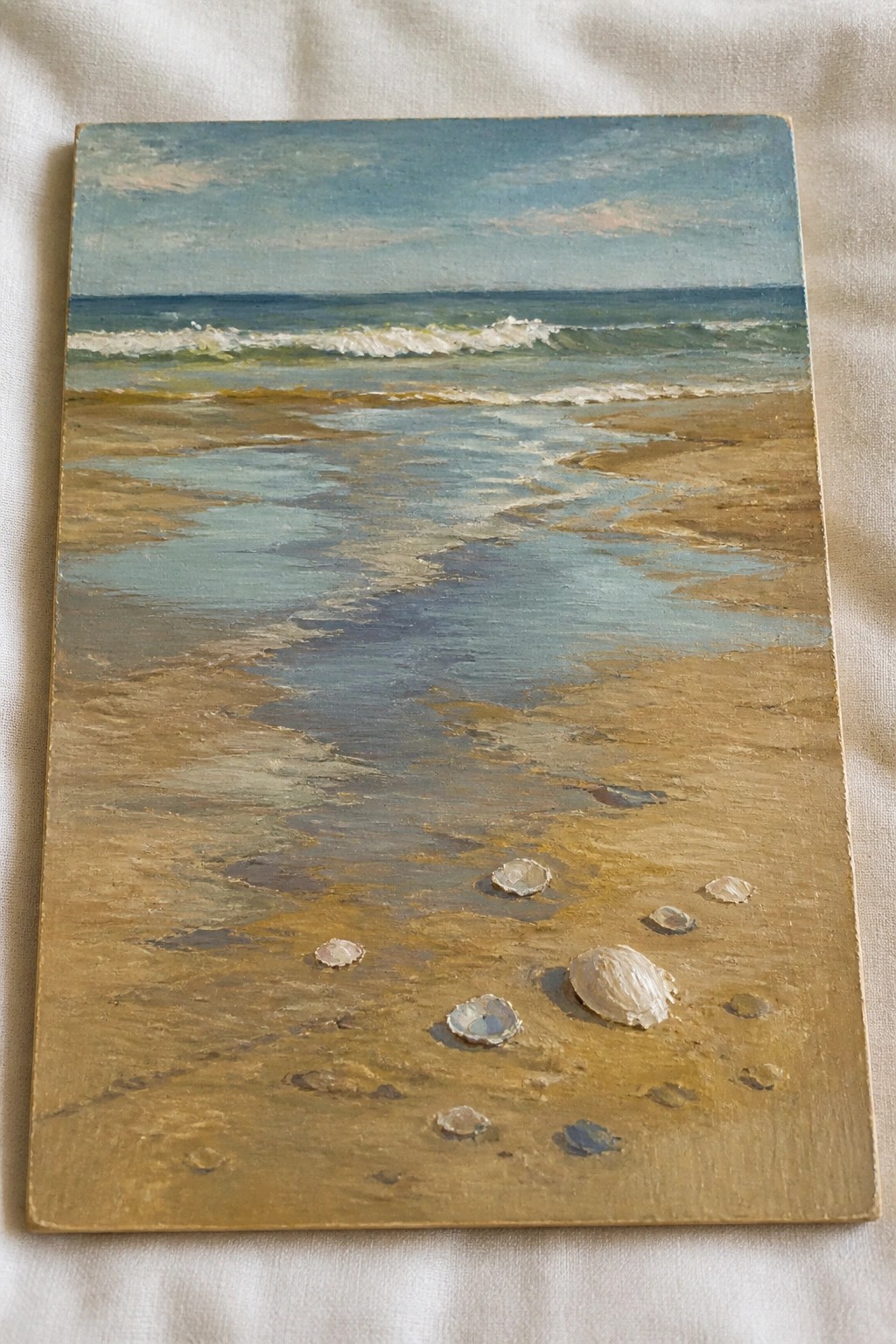 An oil painting of a beach scene showing wet sand, scattered seashells, a shallow water channel, ocean waves, and a blue sky.