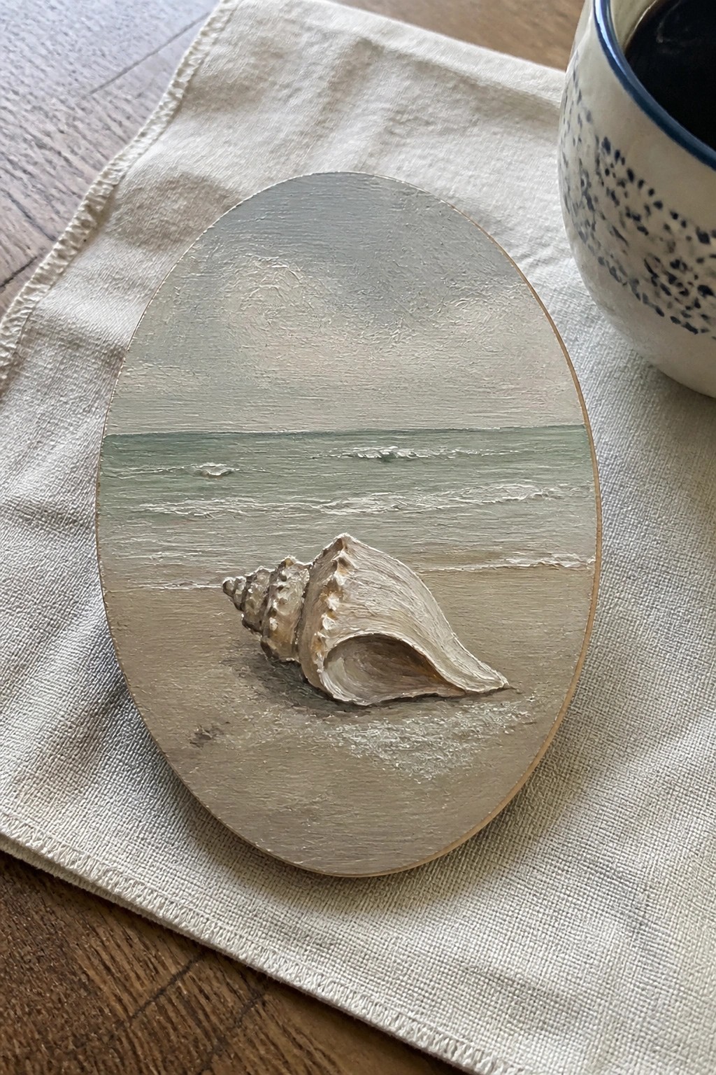 Oval painting of seashell on beach beside blue-speckled mug on linen cloth