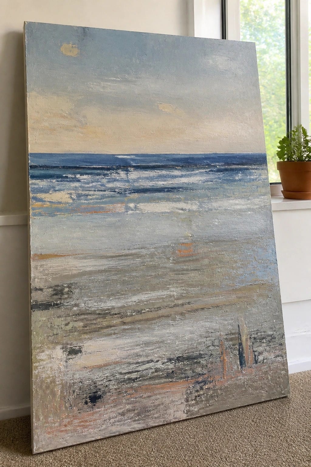Abstract seascape painting in blues and grays leaning against wall by window with plant