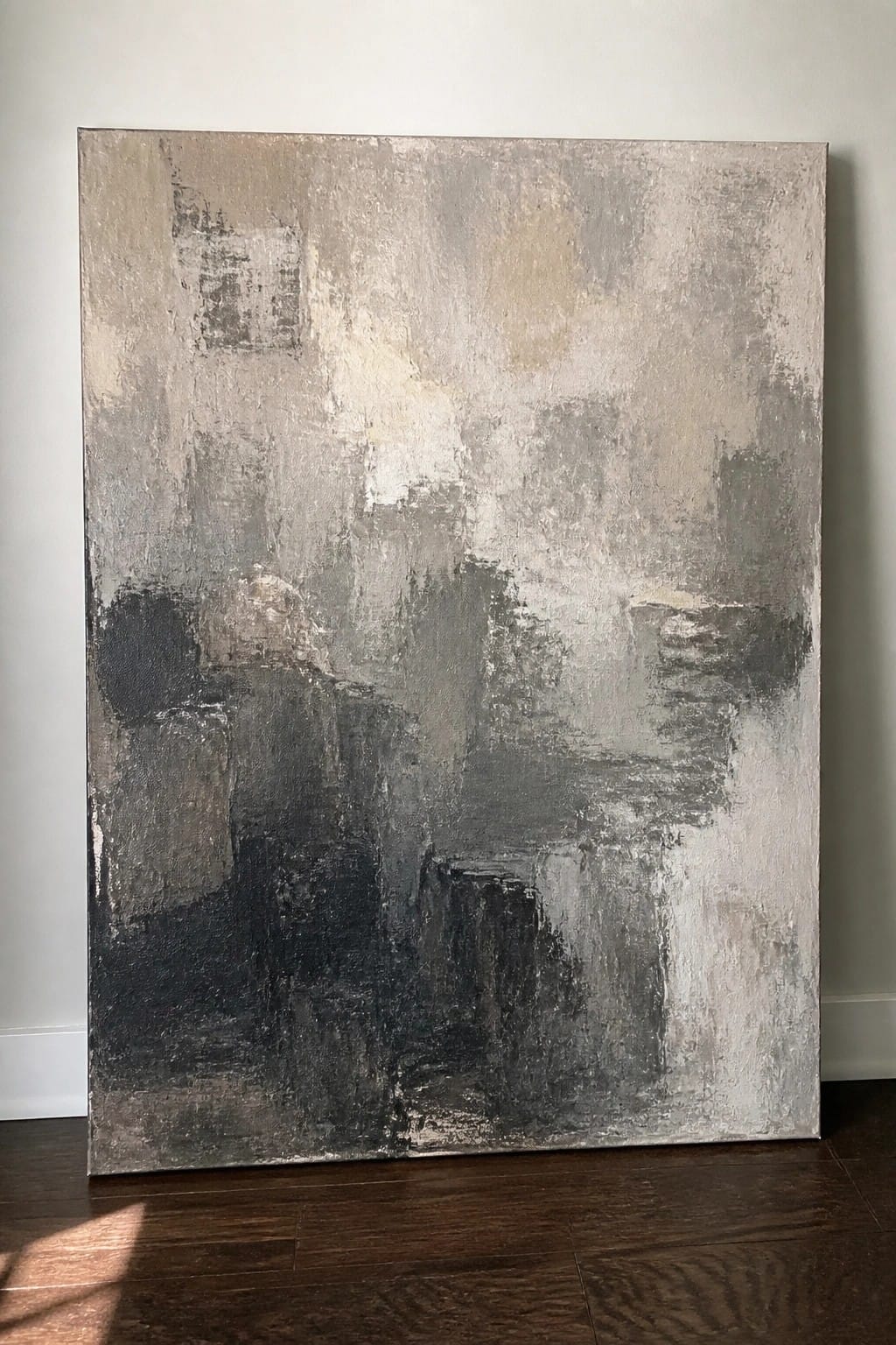 Abstract painting with textured gray and black brushstrokes leaning against white wall on wood floor