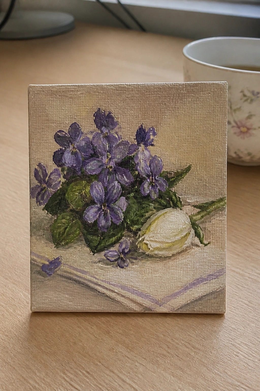 Small canvas painting of purple violets, white bud, and leaves on striped cloth.
