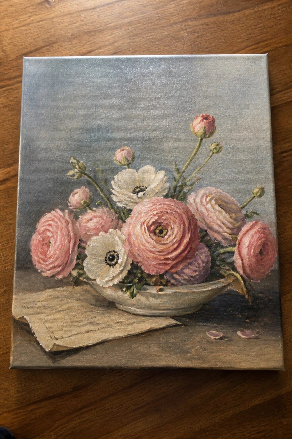 An oil painting of pink and white ranunculus flowers arranged in a shallow white bowl on a wooden surface with a folded paper underneath and a muted gray background.