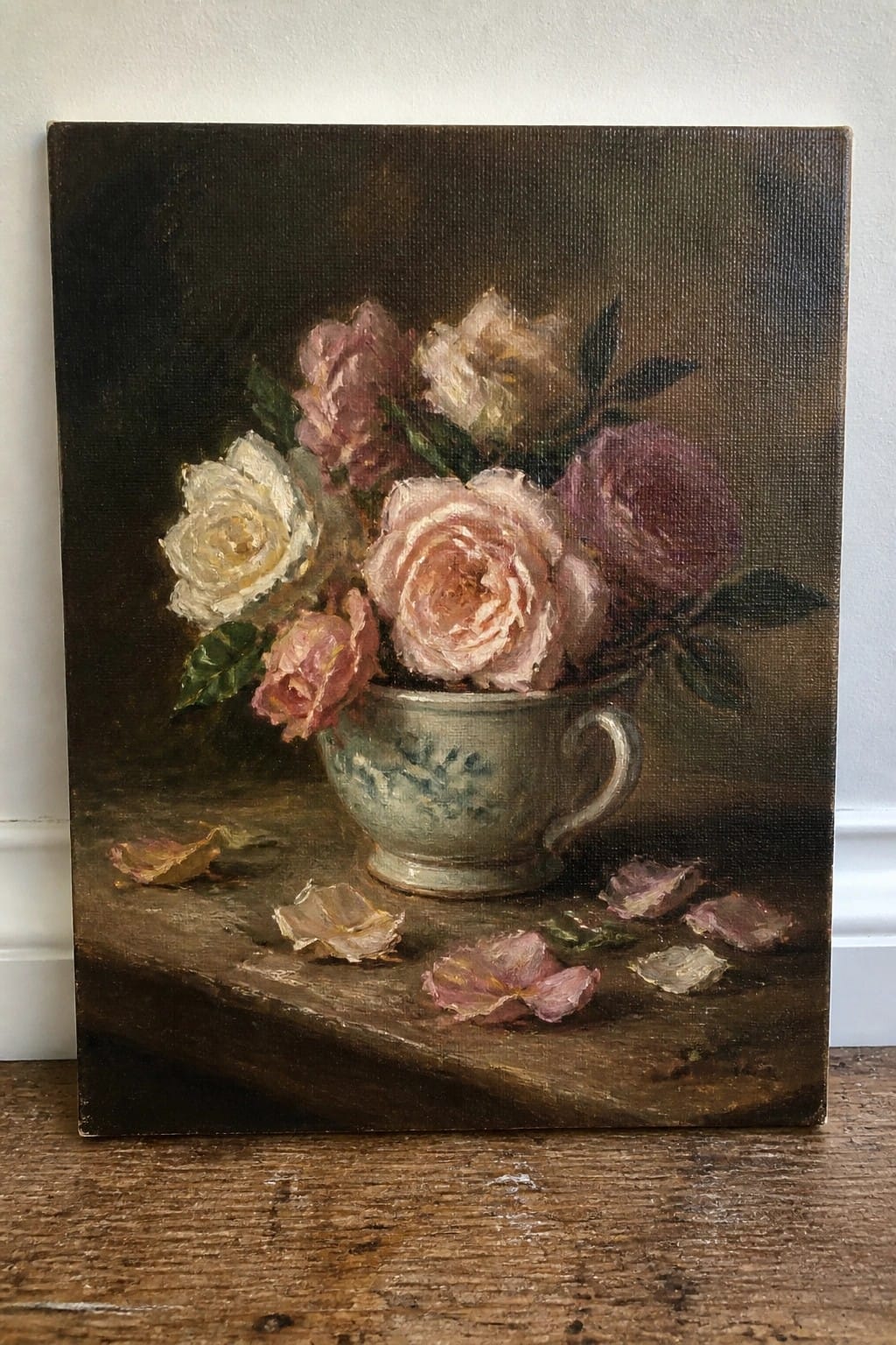Still life painting of pink and white roses in a floral teacup on wood with petals.