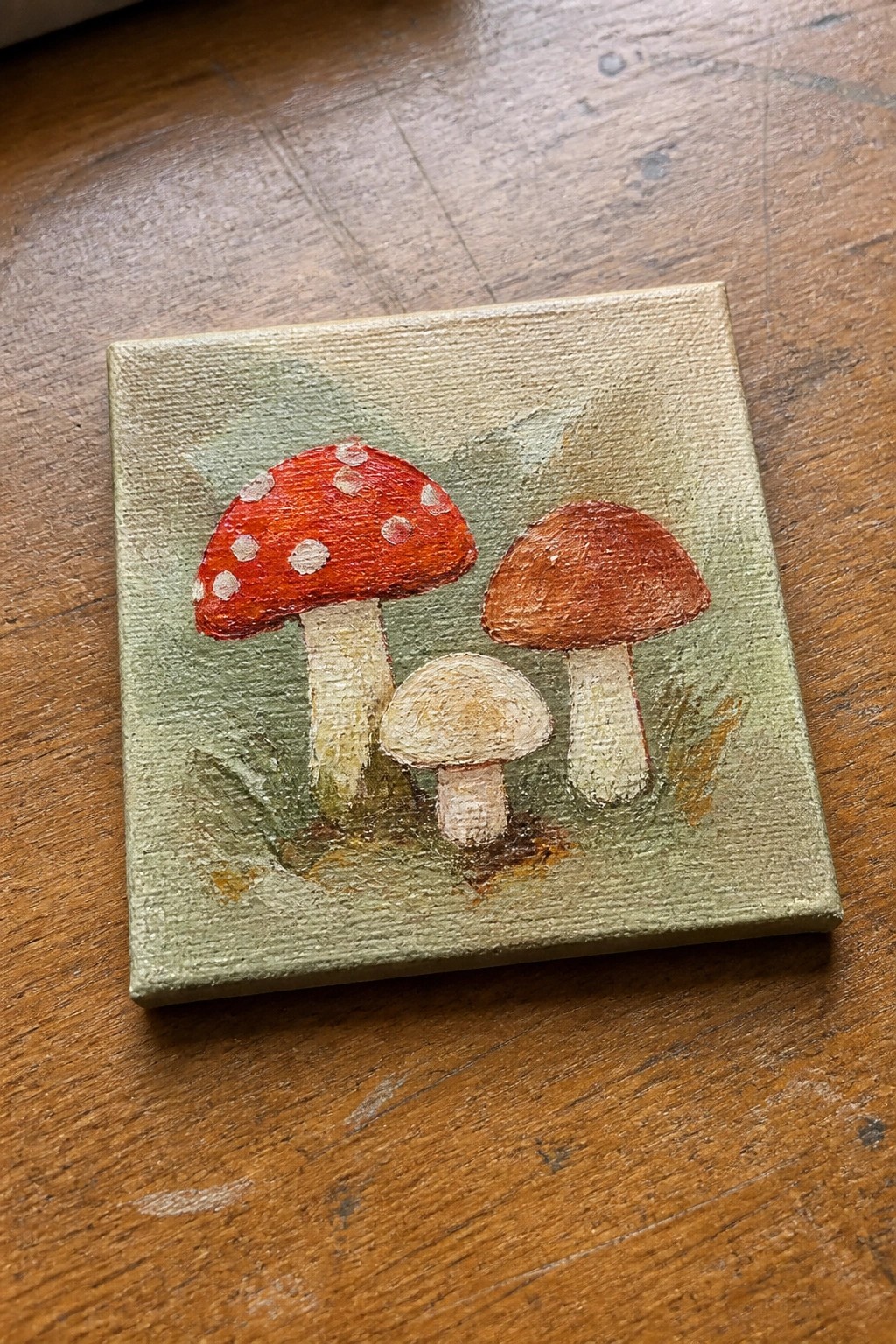 Small canvas painting of three mushrooms: red spotted, brown, and beige on green.