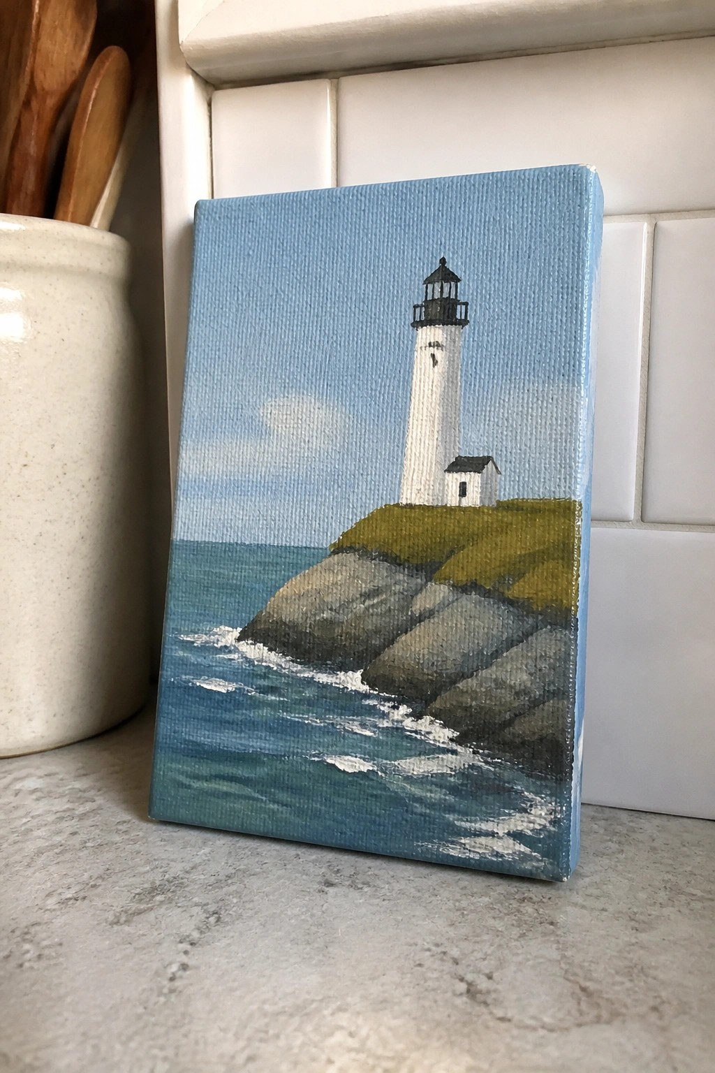 Small painting of white lighthouse on rocky cliffs by blue ocean waves.