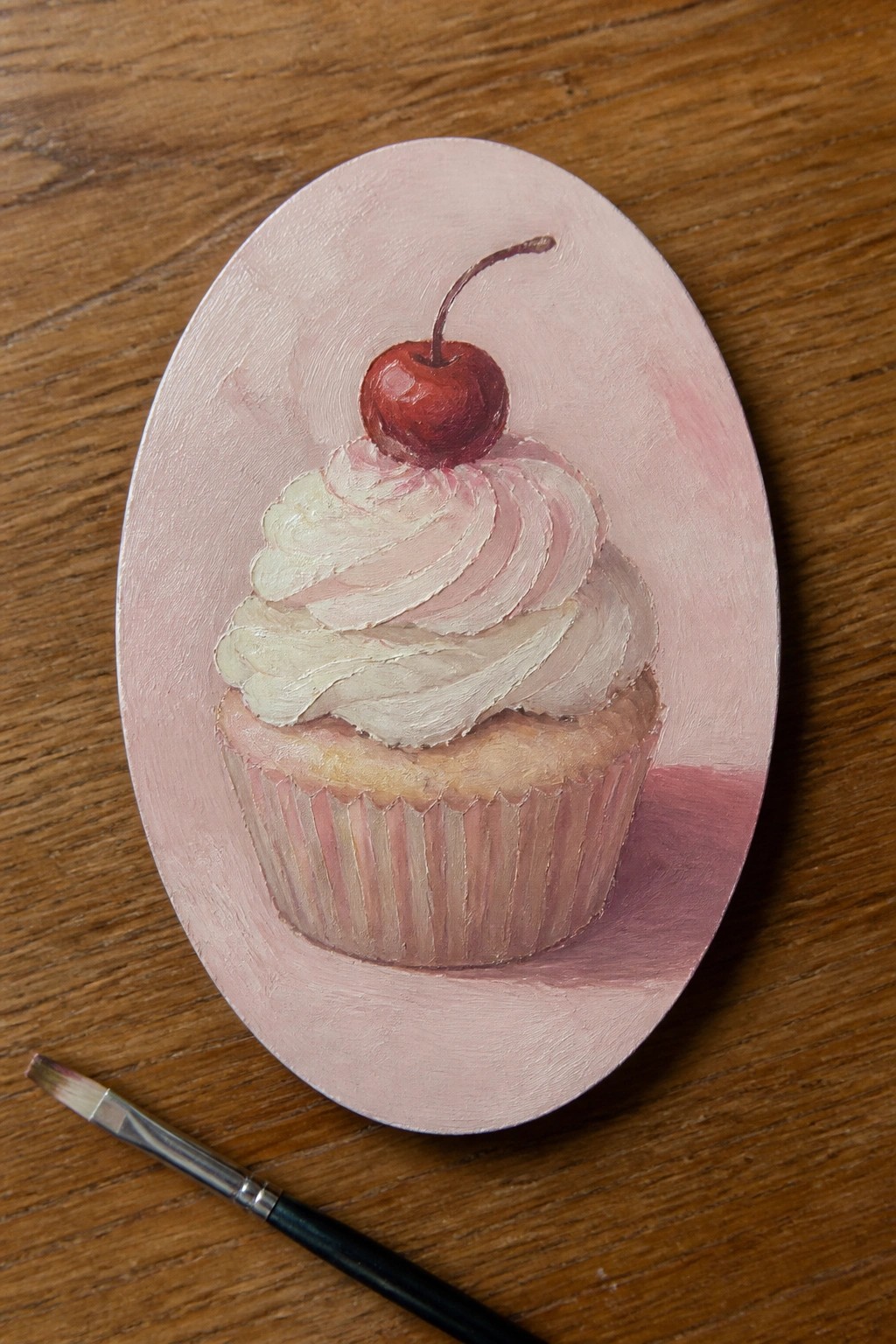 Realistic painting of cherry-topped cupcake on pink oval canvas beside paintbrush.