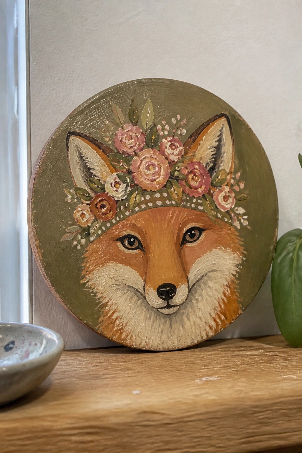 A circular oil painting of a fox face wearing a crown of pink and rust roses with green leaves on an olive green background, resting on a wooden shelf.