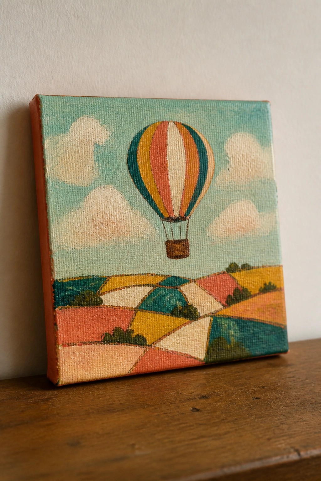 Colorful striped hot air balloon over patchwork fields on small canvas resting on wood.