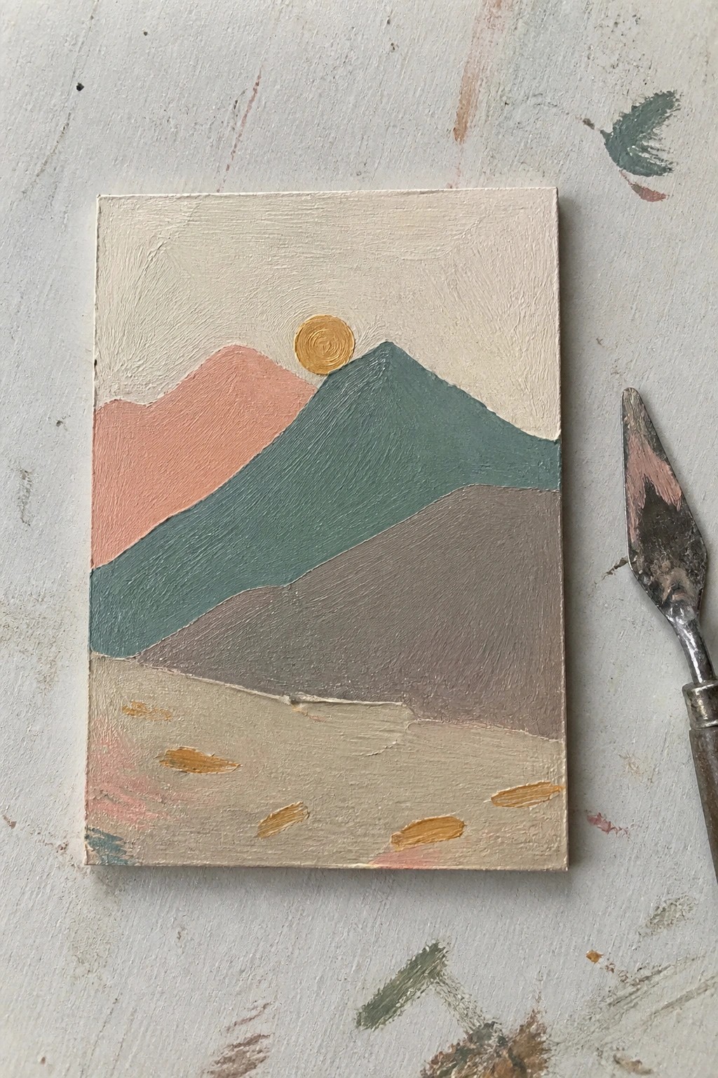 Abstract landscape painting of pastel mountains and golden sun on canvas beside palette knife.