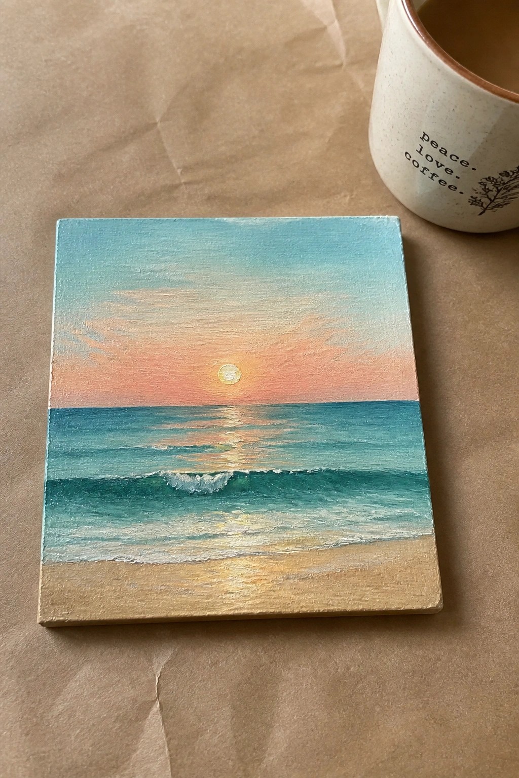 Mini canvas sunset seascape painting beside white peace love coffee mug on brown paper.