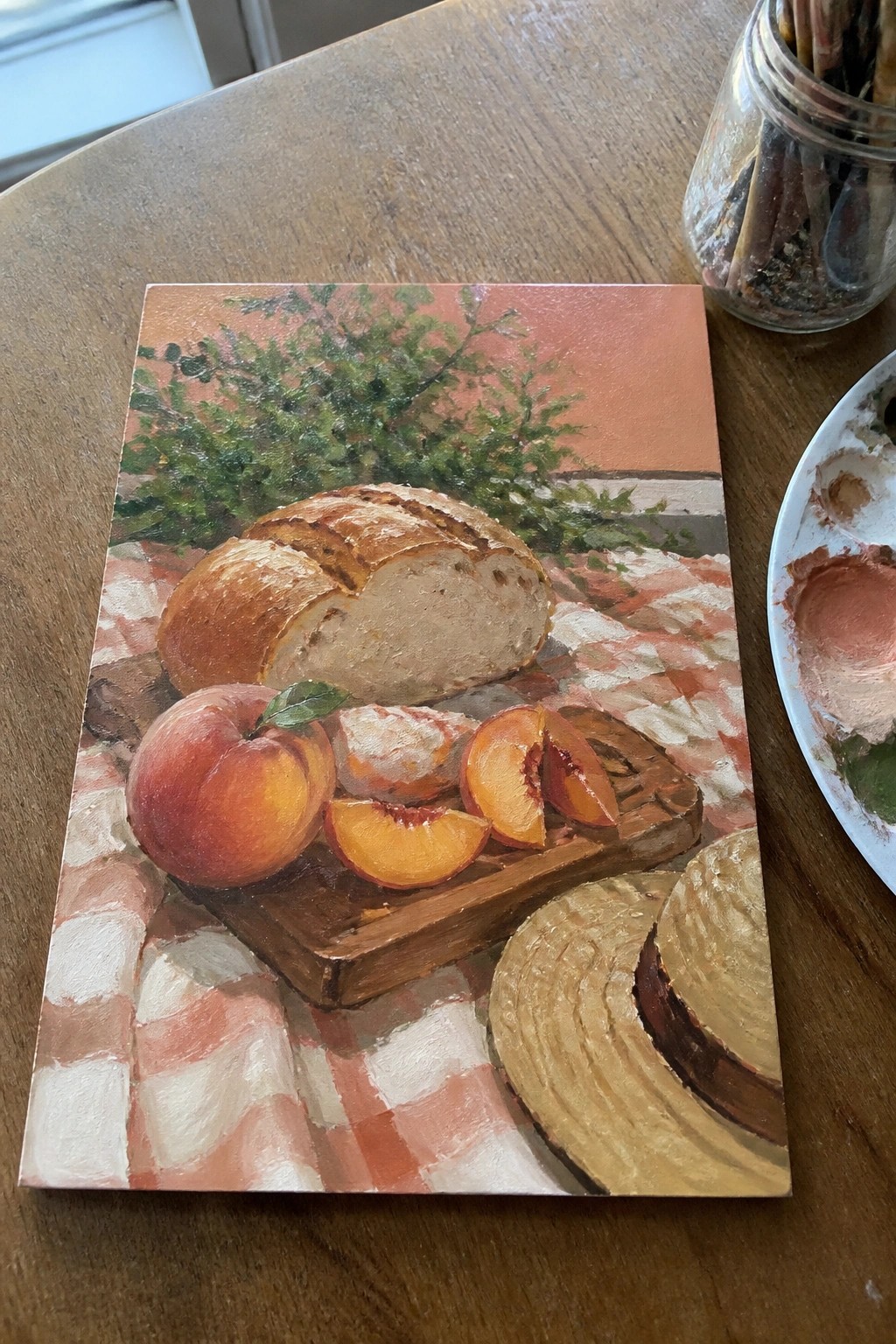 Still life painting of bread, peaches, straw hat on red checkered cloth with foliage.