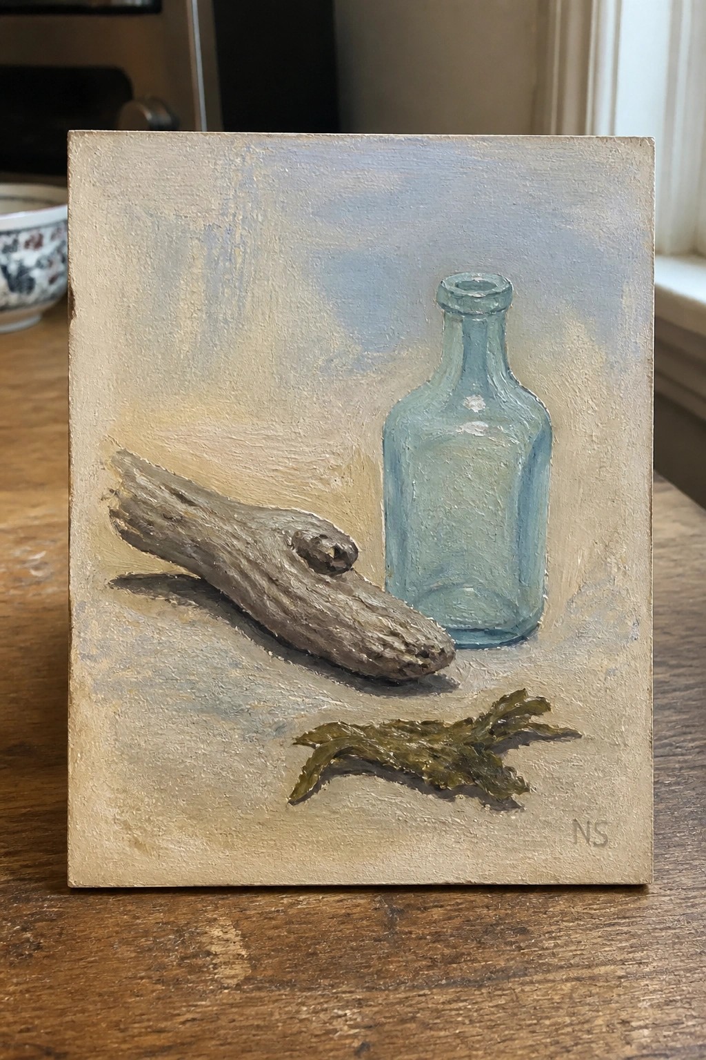 Still life painting of light blue glass bottle, driftwood, and green seaweed on beige.