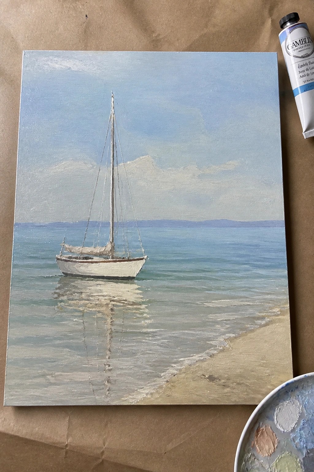Oil painting of white sailboat on blue water by sandy beach, with paint tube and palette nearby.