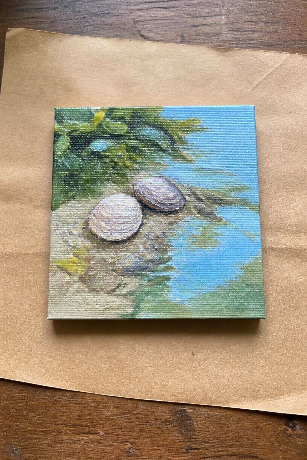 Small square painting of two seashells on sandy shore by blue water with green foliage