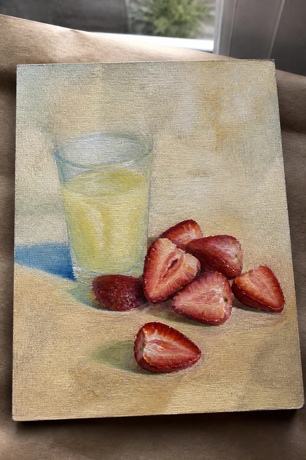 Still-life painting of yellow juice glass beside halved red strawberries on beige surface.