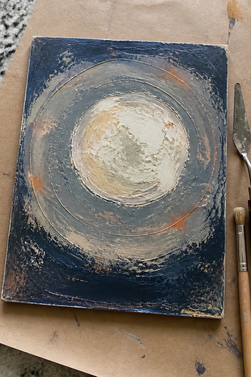 Abstract painting with concentric blue circles, textured beige center, beside palette knife and brush.