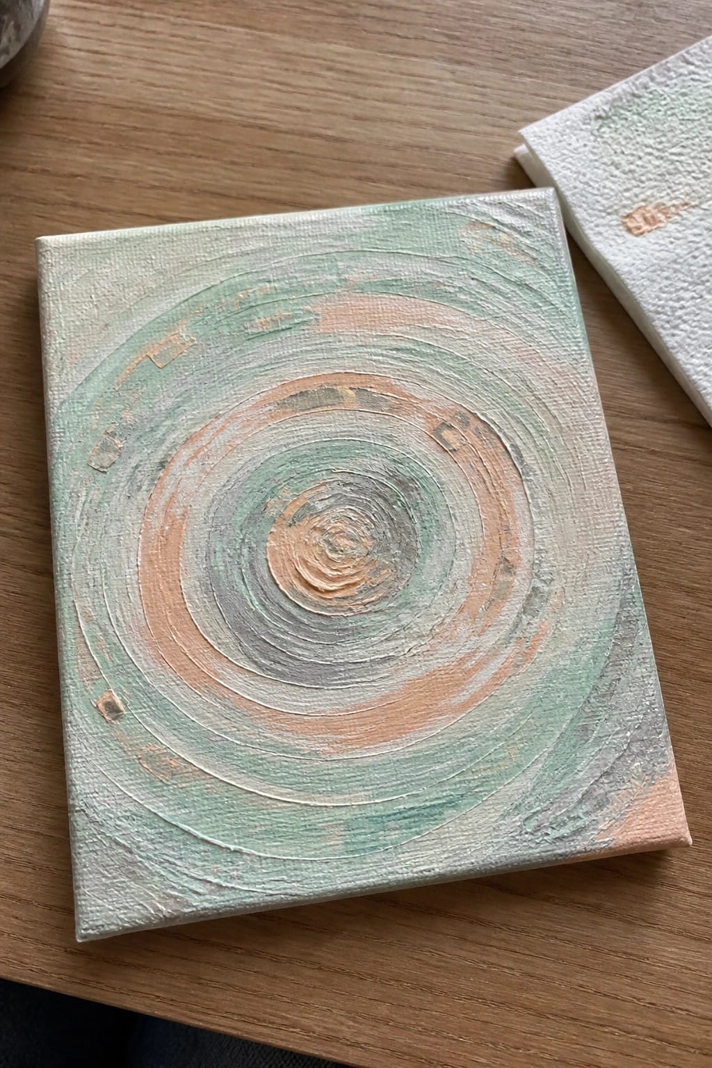 Square canvas with swirling green and peach concentric circles on wood beside napkin
