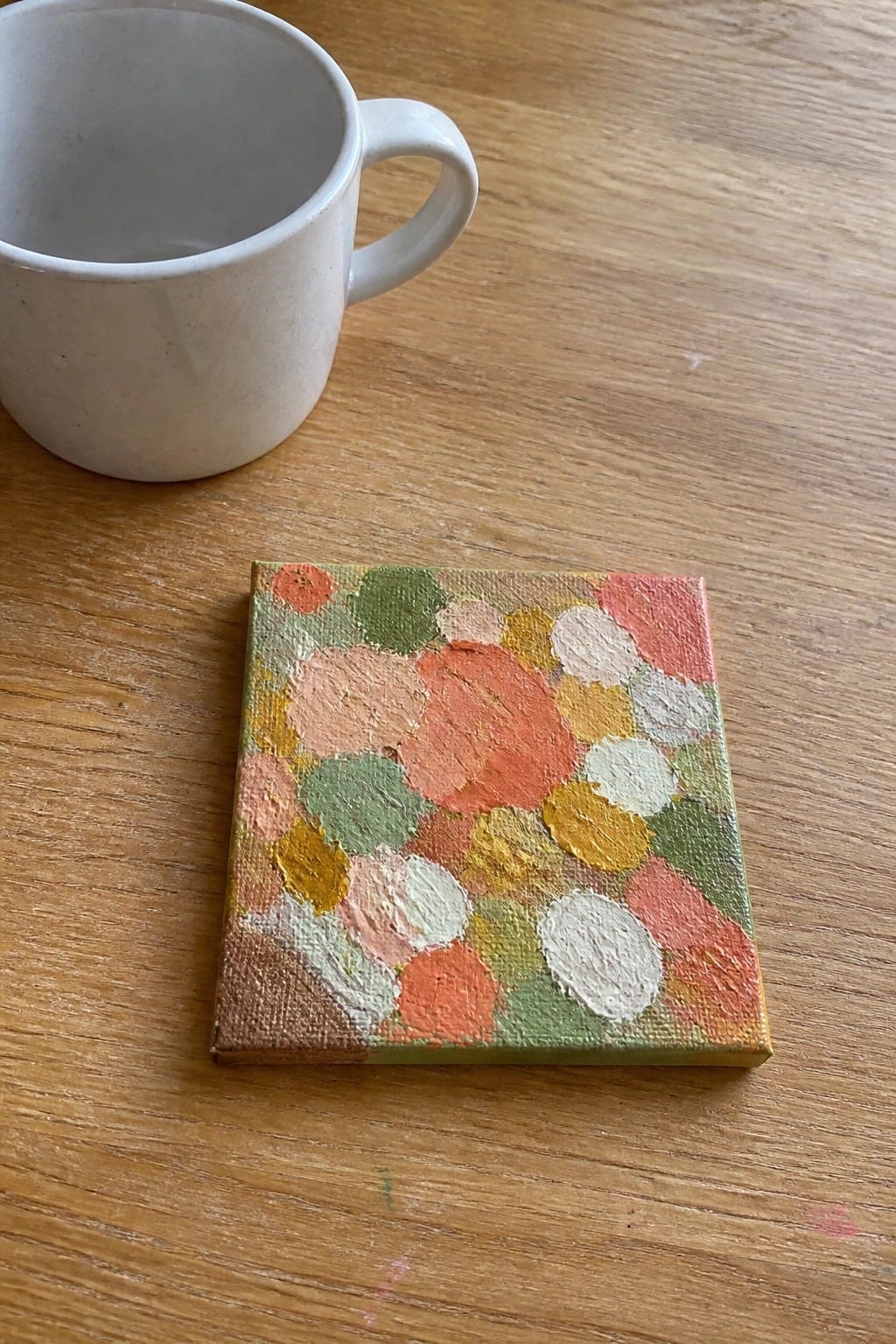 A small square canvas covered in overlapping painted circles in coral, peach, mustard, sage, and cream rests on a wooden table beside a white mug.