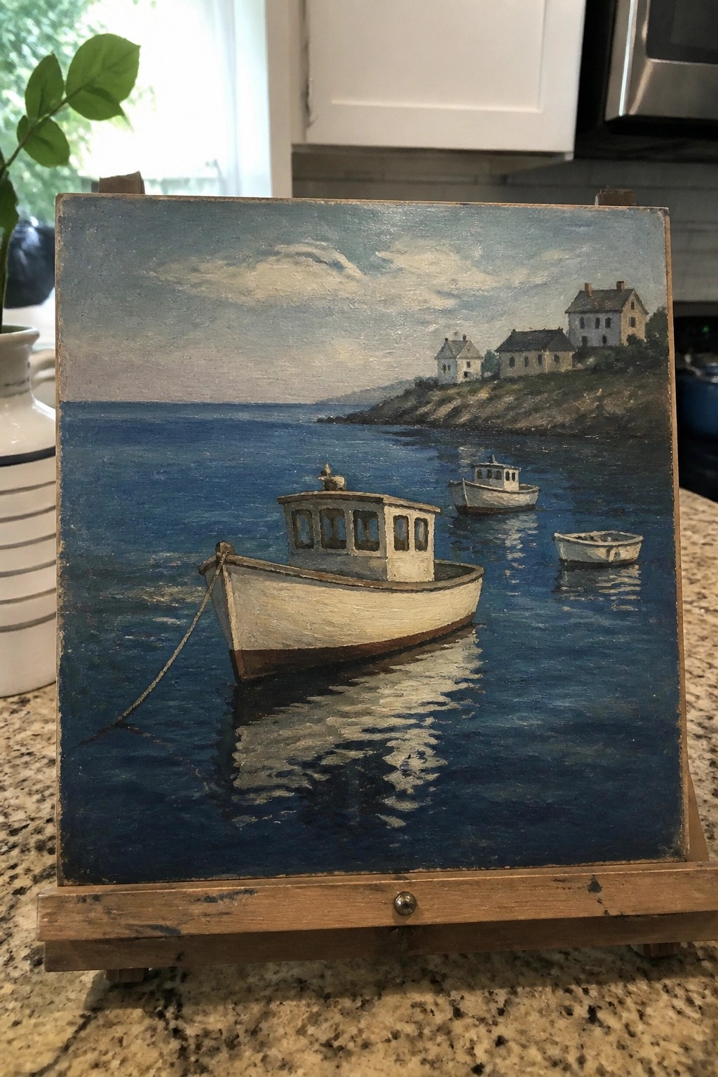 Realistic oil painting on easel of three boats on blue water near hillside houses