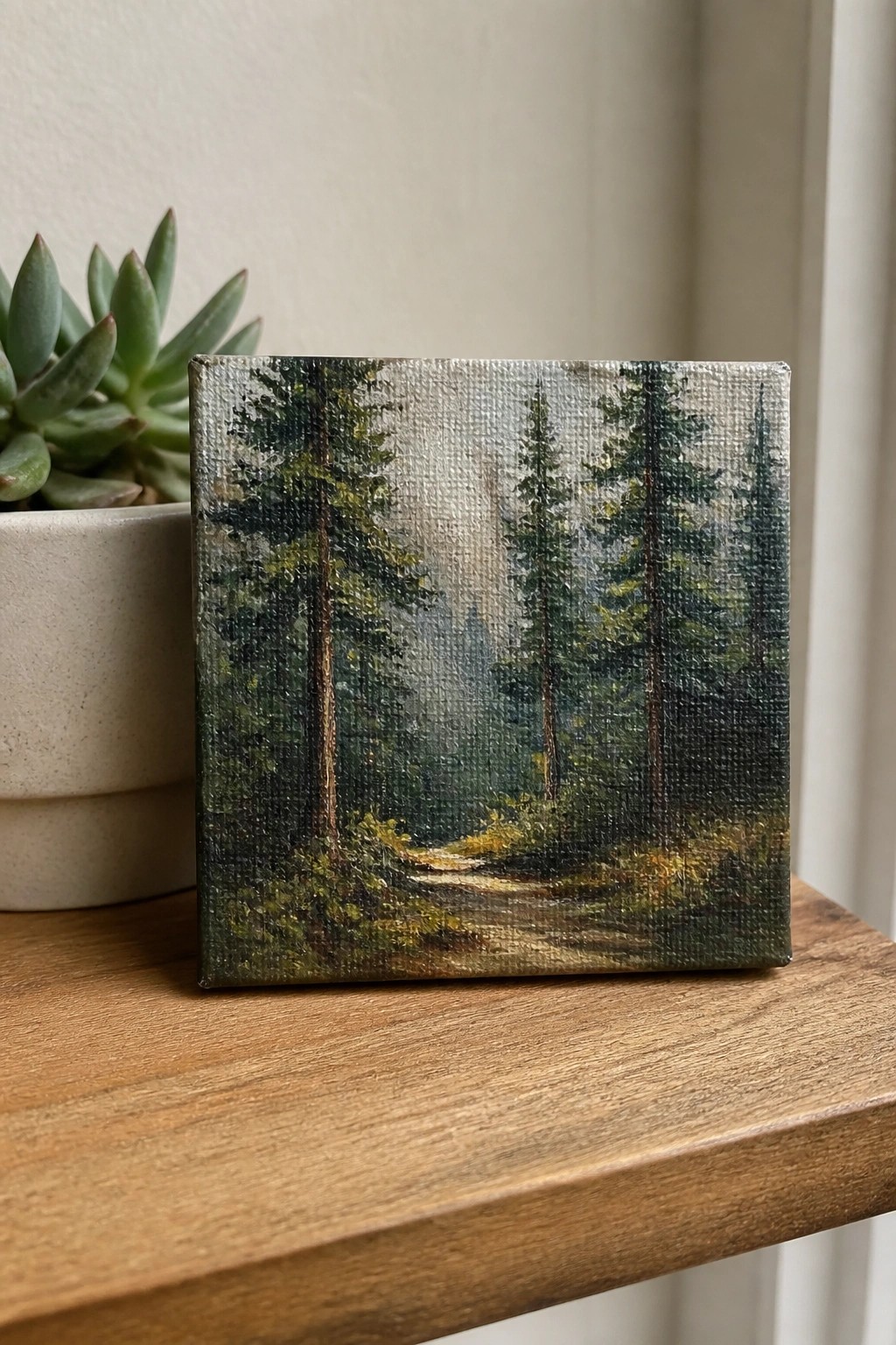 Small square painting of misty forest path with tall trees beside potted succulent on wood.