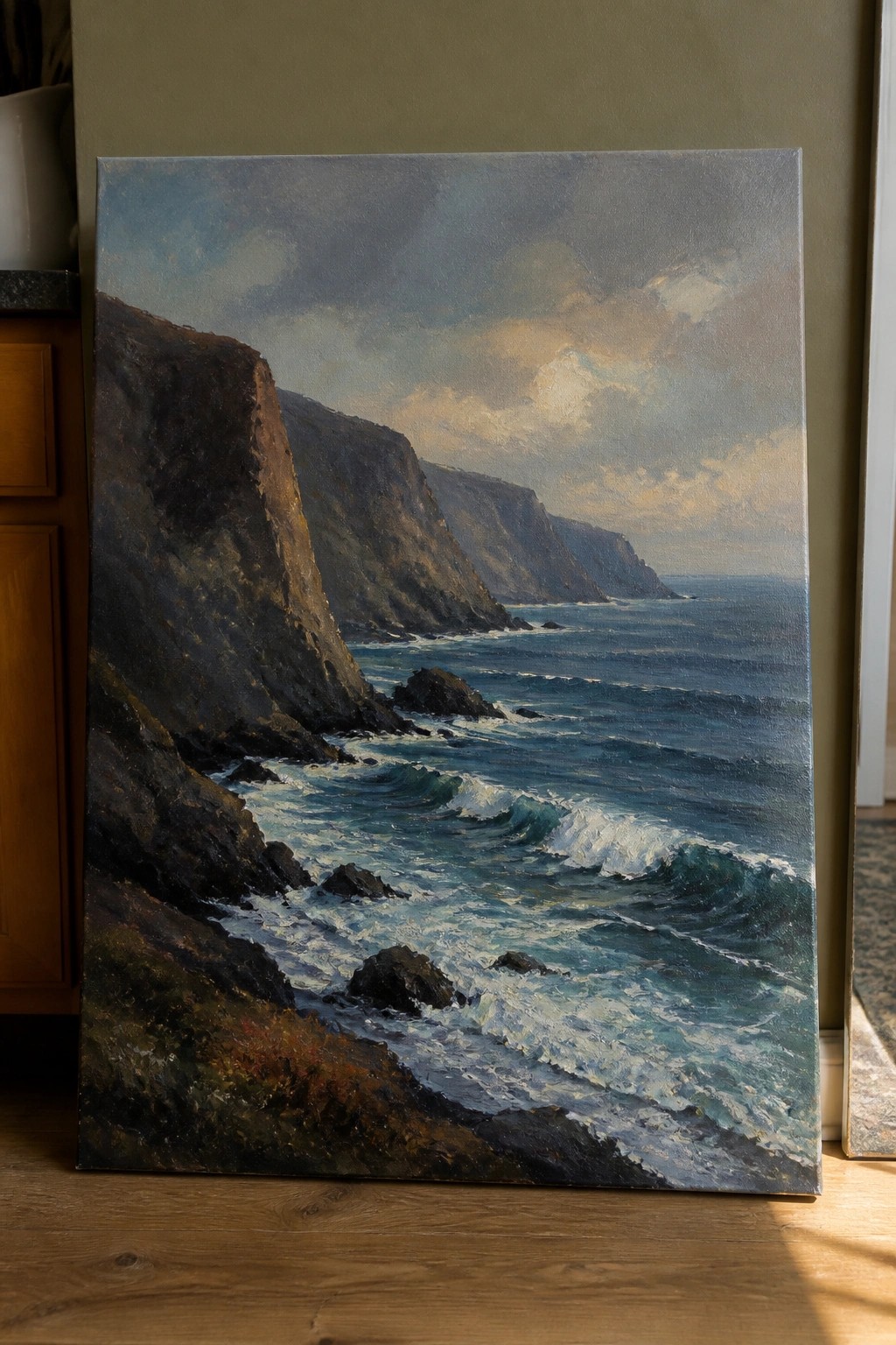 Large oil painting of rugged cliffs and crashing blue waves propped on wooden floor