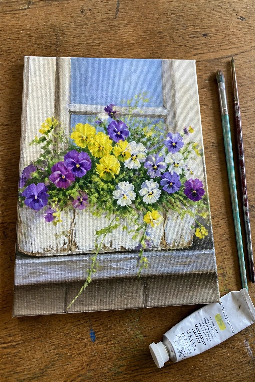 Colorful pansy painting on canvas with paintbrushes and yellow paint tube on wood