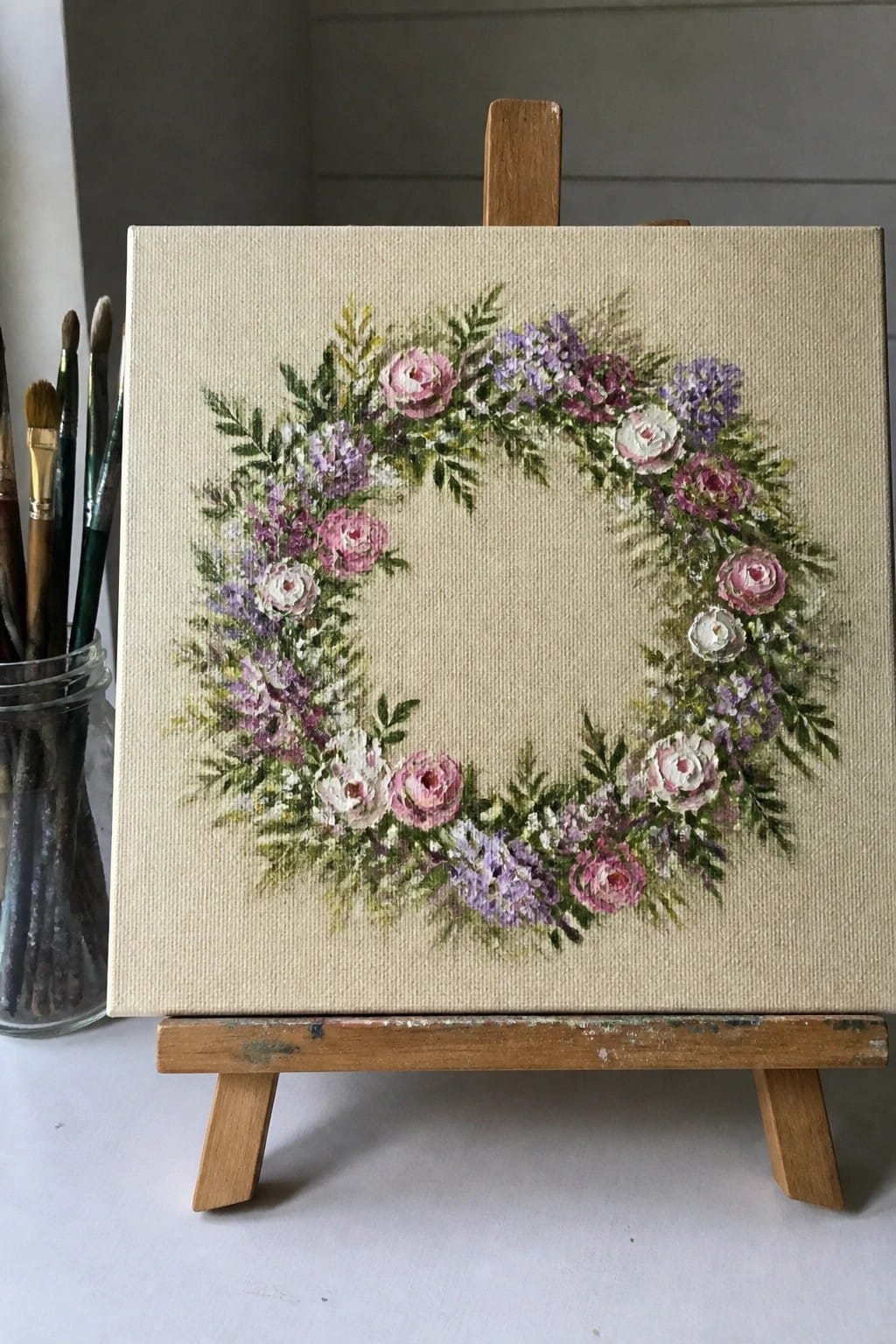 Canvas on easel with pink rose and purple floral wreath painting beside brushes